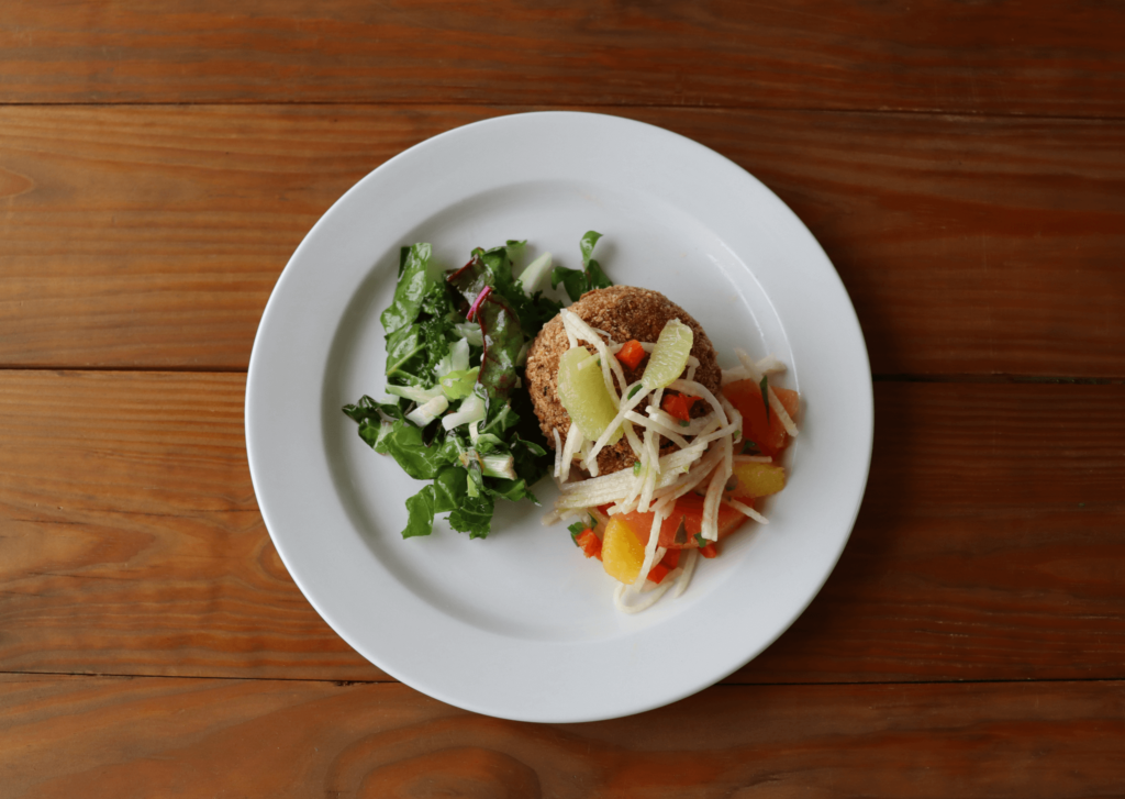 A plate with a quinoa patty topped with sliced vegetables, accompanied by a side of fresh leafy greens on a wooden table.