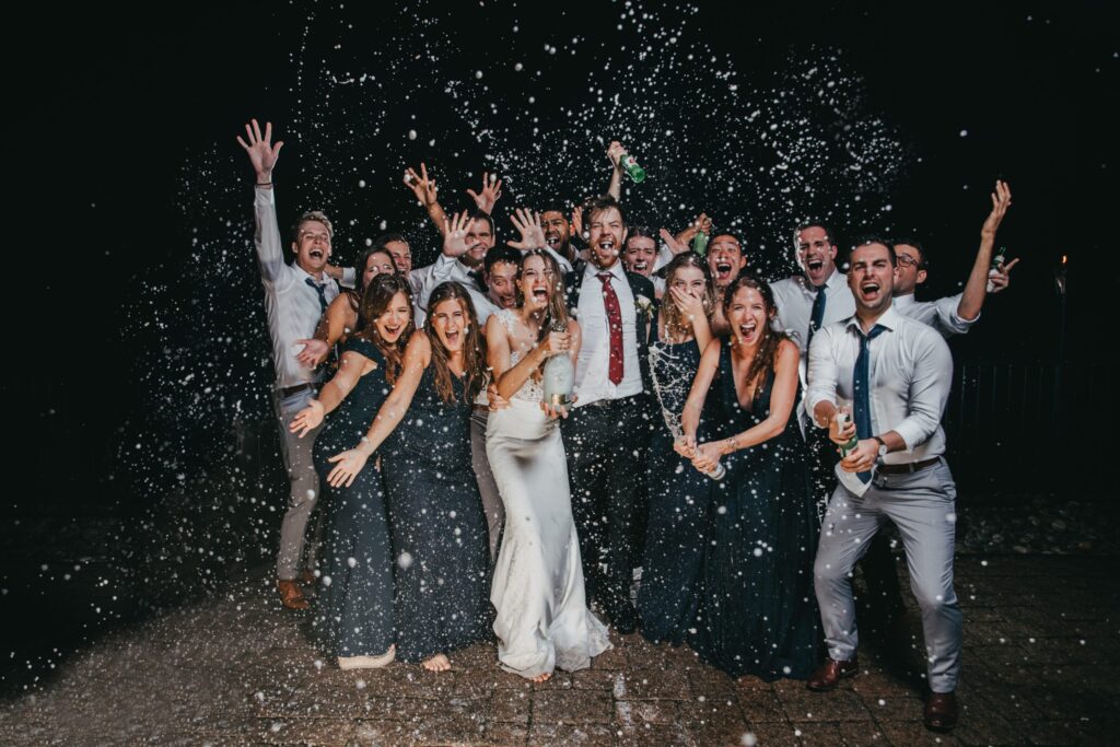 A group of people celebrates energetically, with champagne spraying around, at what appears to be a nighttime event or party.