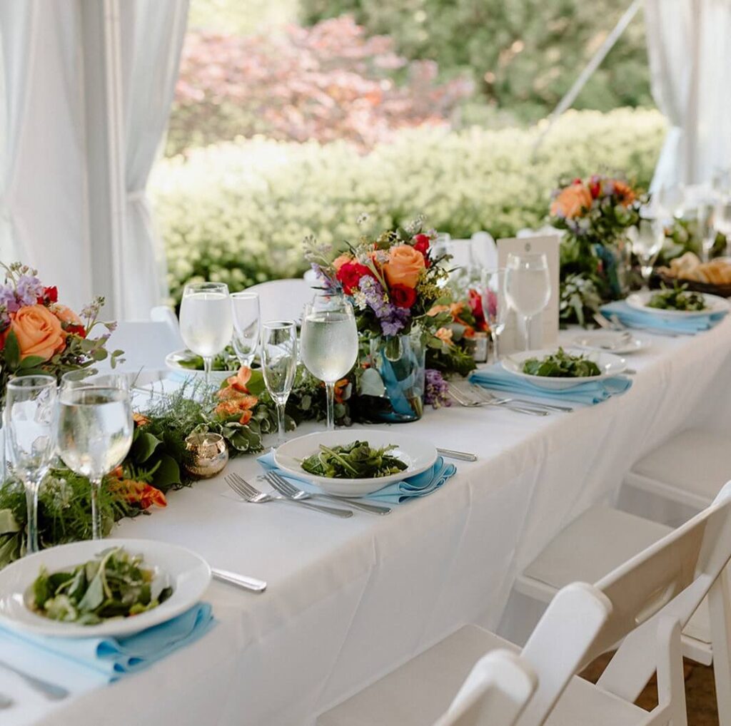 Elegant outdoor table setting with white tablecloth, flowers, glassware, and plates of salad. Bright floral centerpieces and greenery in the background.