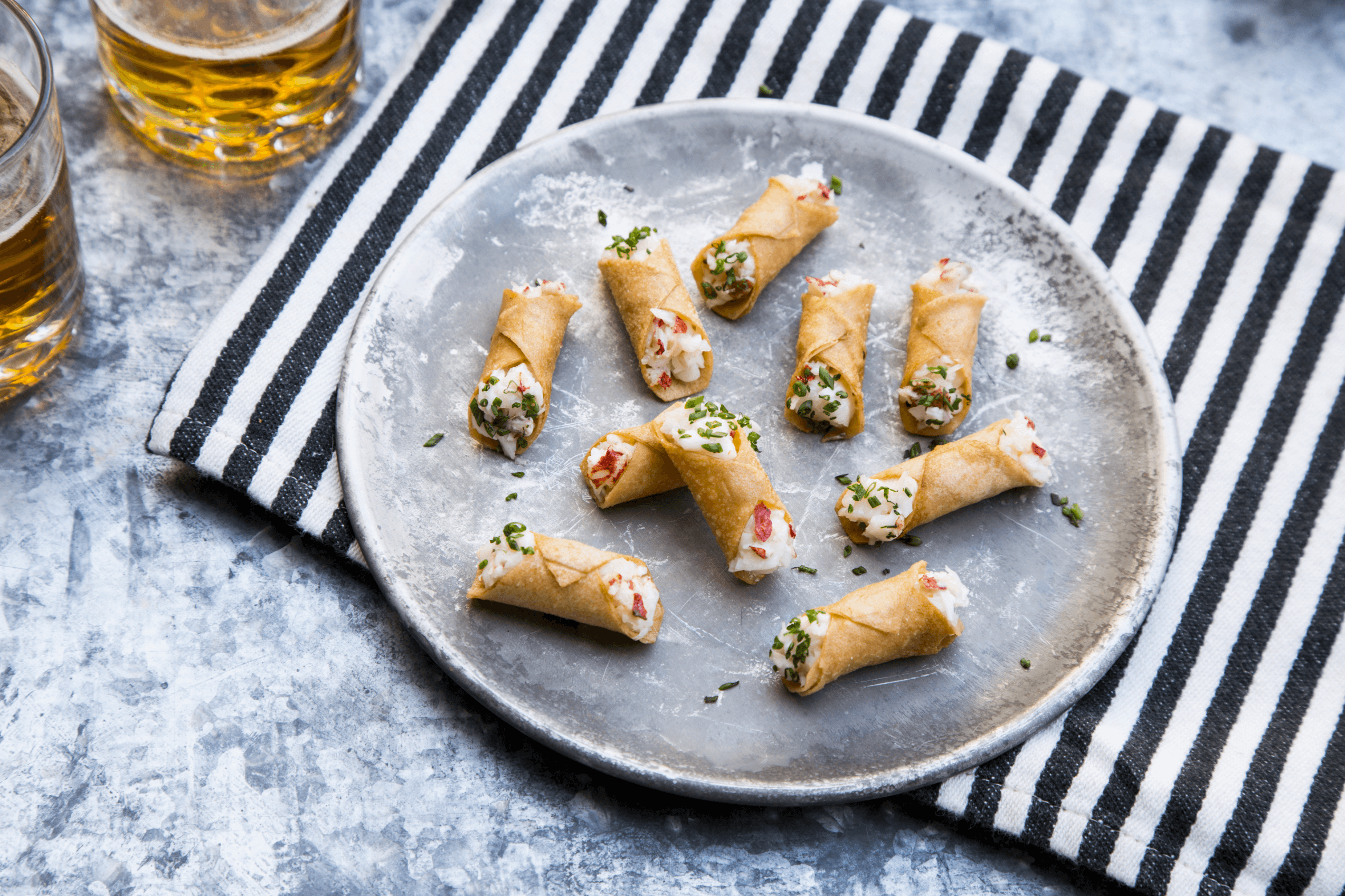 Plate of mini cannoli filled with cream cheese and chives on a striped cloth beside two glasses of beer.