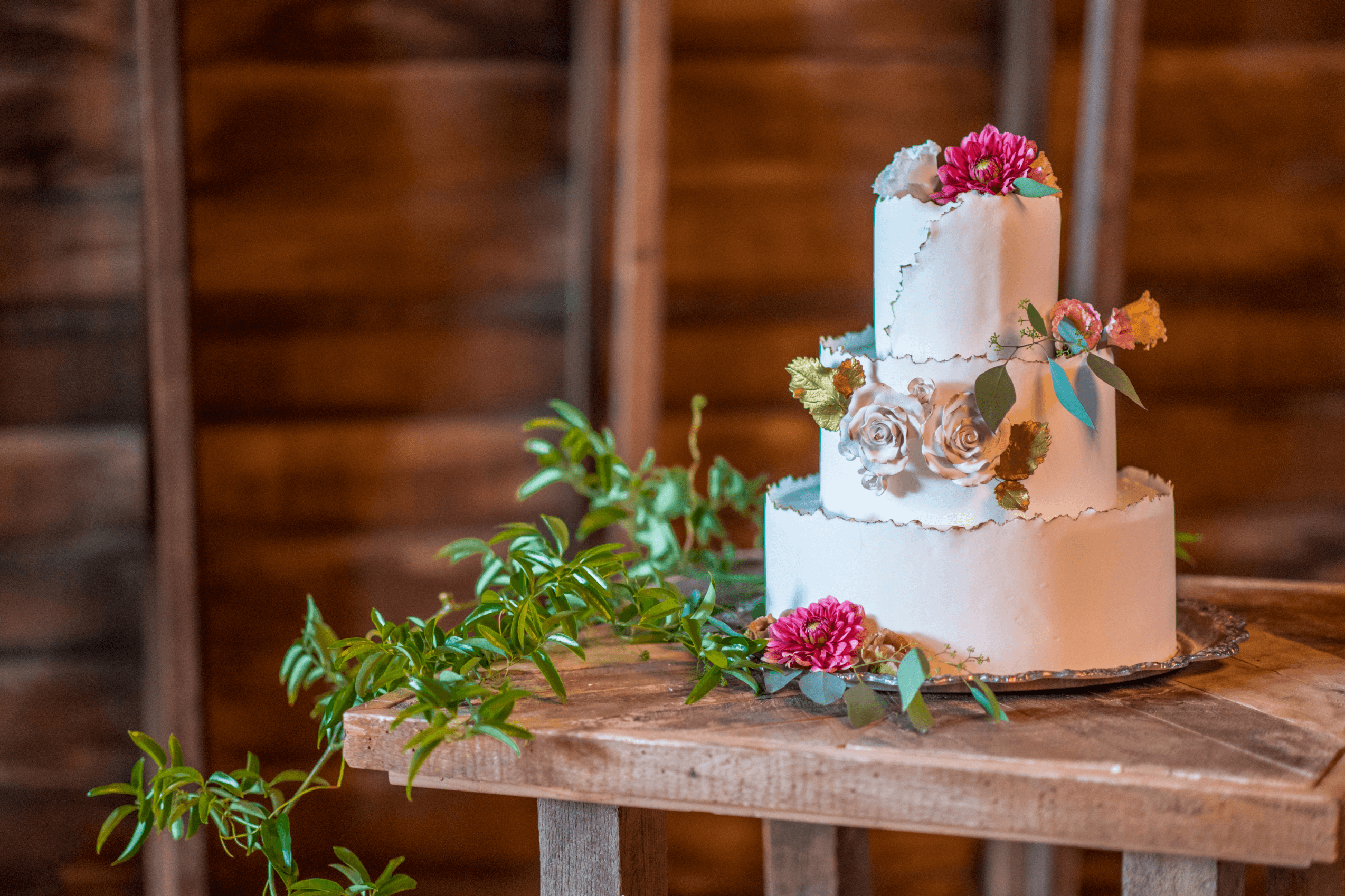 Three-tier white cake with floral decorations and greenery on a wooden table against a wooden wall.