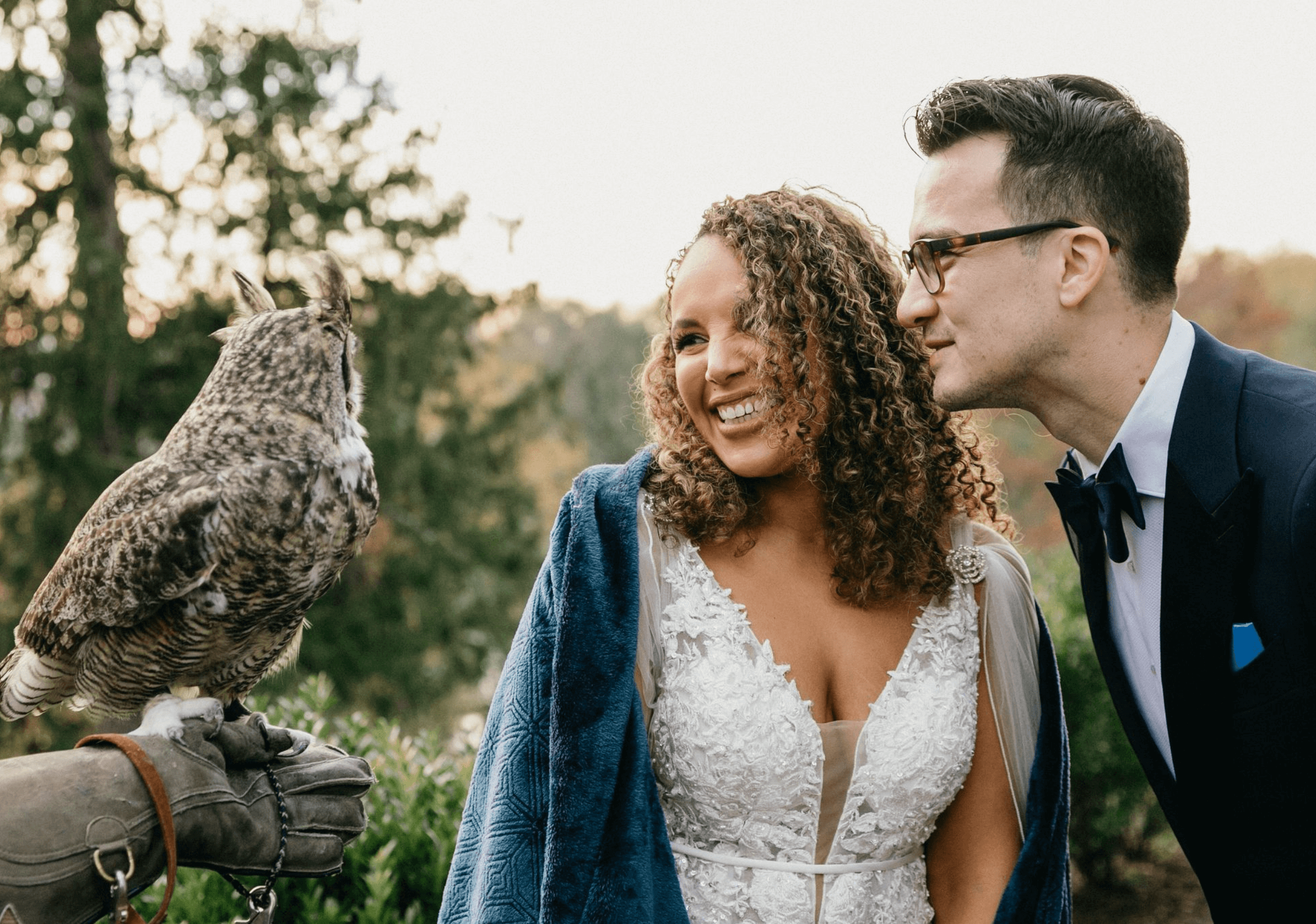 A bride and groom smile at an owl perched on a gloved hand. The bride wears a lace dress and blue shawl; the groom is in a tuxedo. Trees are in the background.