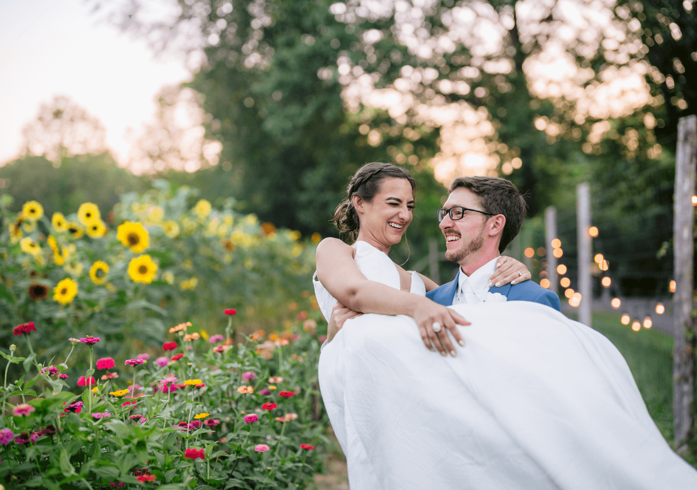 A groom in a blue suit carries a bride in a white dress through a garden path lined with sunflowers and string lights. Both are smiling, with trees in the background.