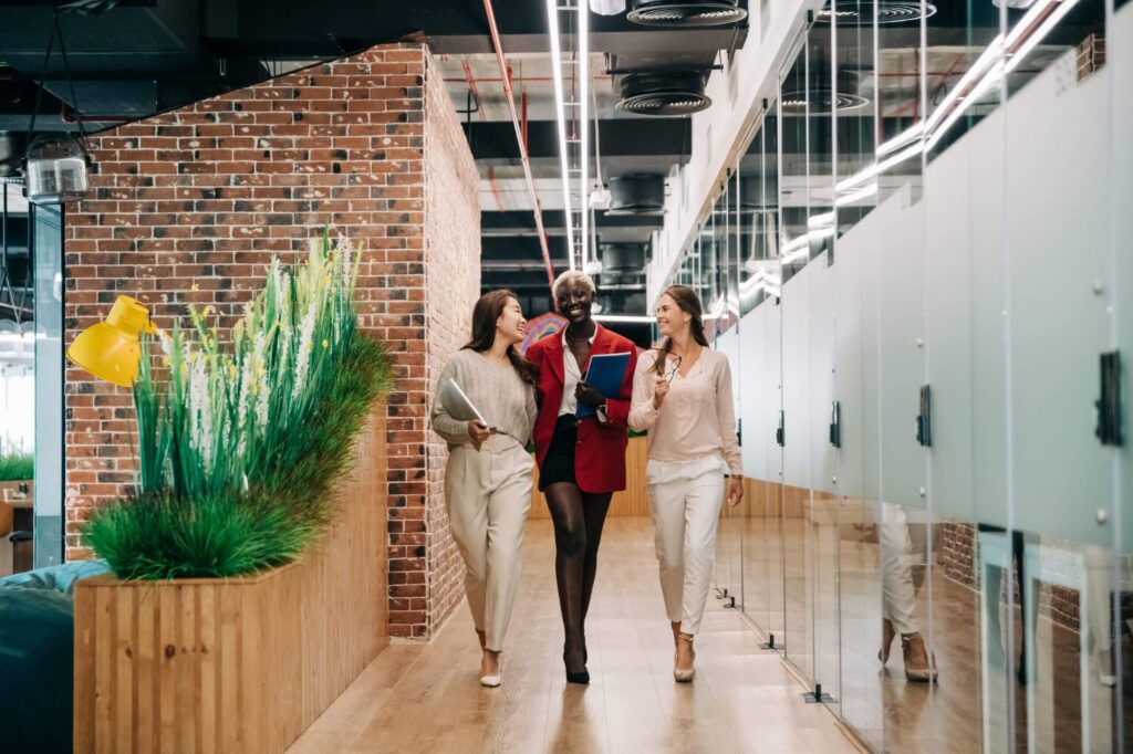 Three people walk together down a modern office hallway, smiling and carrying folders. The corridor features brick walls and decorative plants.