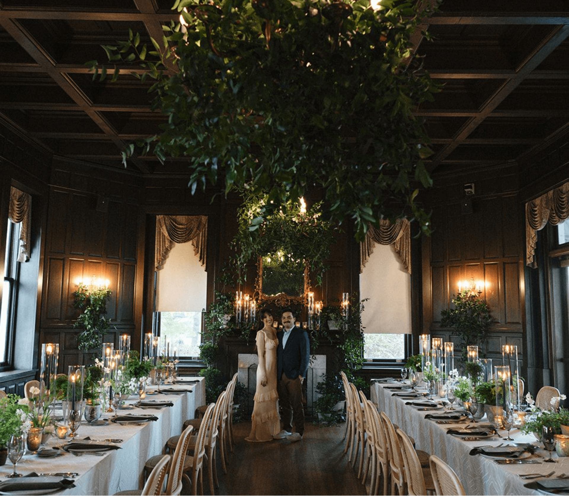 A couple stands in a dimly lit, elegantly decorated dining hall with long tables, greenery, and candles.