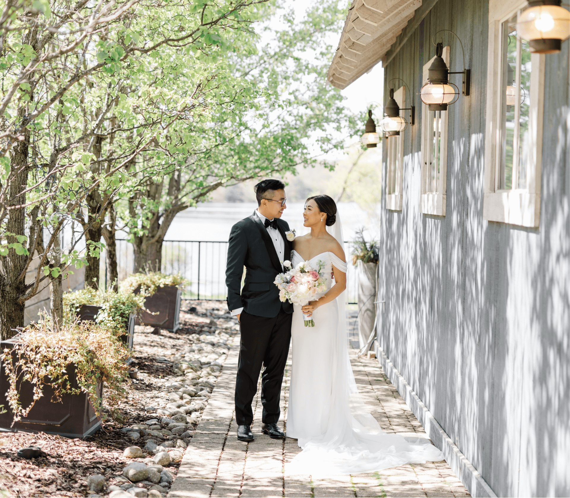 A bride and groom stand together on a stone path near a building, surrounded by trees. The bride holds a bouquet of flowers.