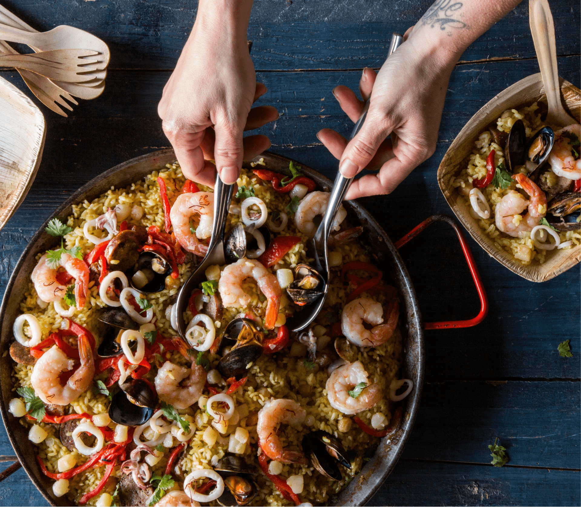 Two hands serve seafood paella from a large pan on a dark wooden table, featuring shrimp, mussels, and slices of red pepper, with a small portion in a square dish.