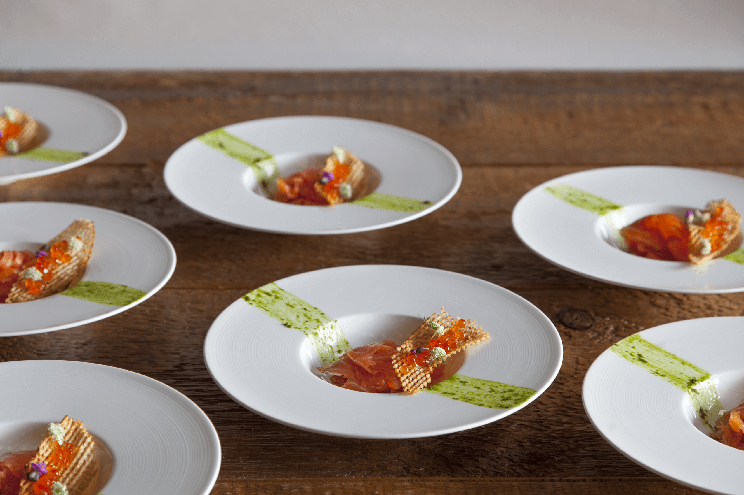 White plates on a wooden table, each with a small serving of salmon, topped with garnishes, and a green sauce stripe on the side.