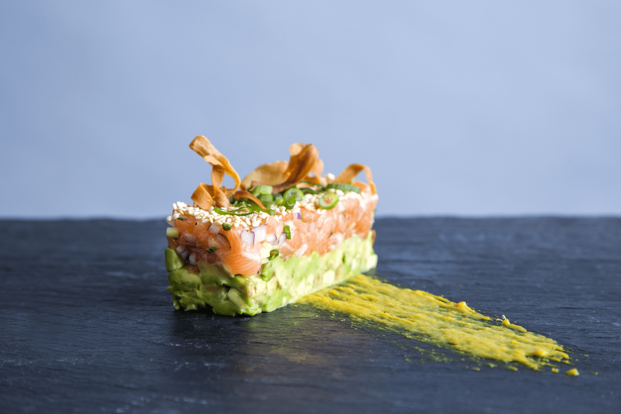 A rectangular stack of diced avocado and salmon topped with sesame seeds and garnishes, on a dark slate surface with a smear of yellow sauce.