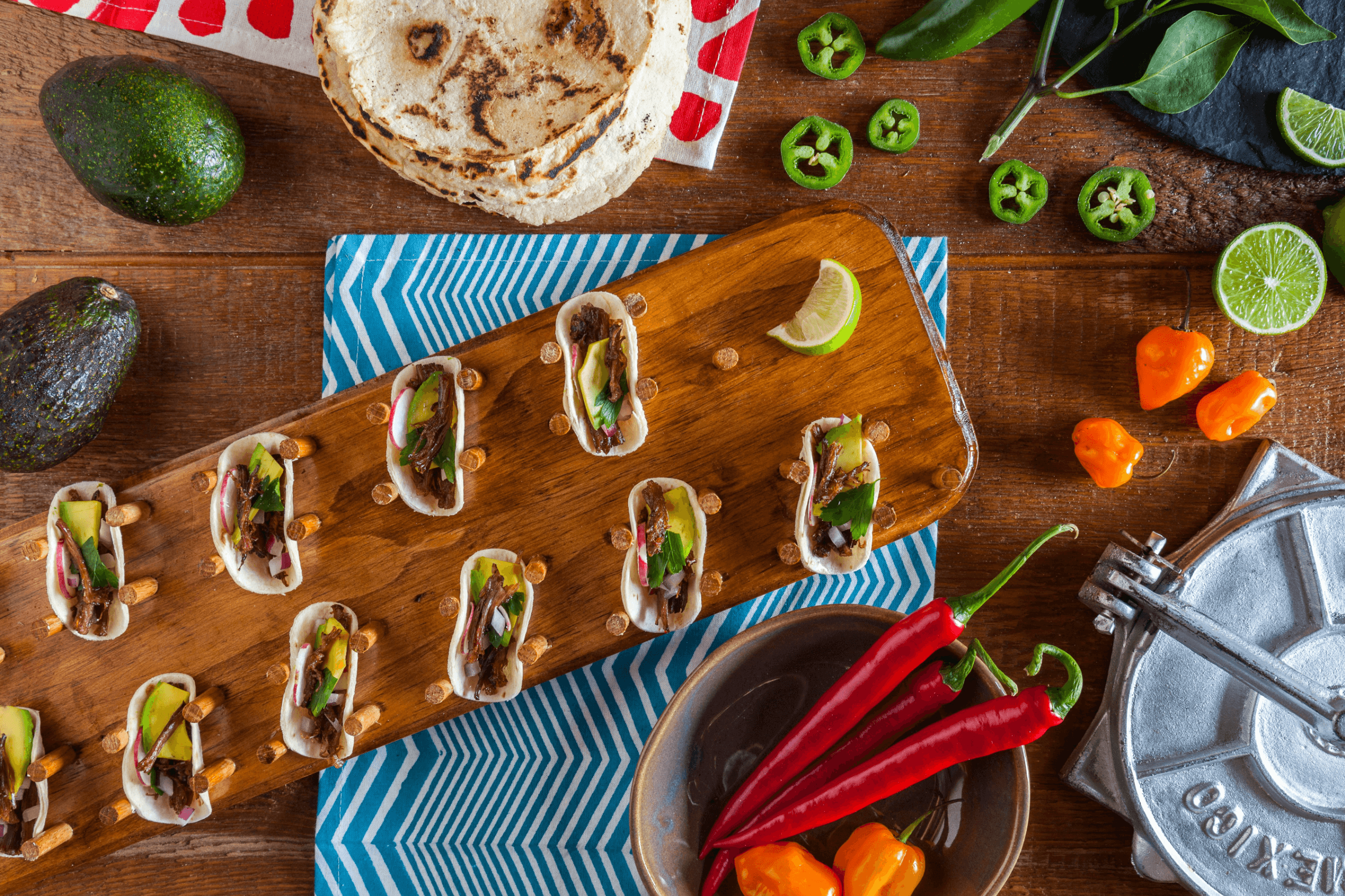 Overhead view of small tacos on a wooden board, surrounded by avocados, red chilies, sliced jalapeños, limes, tortillas, and orange peppers on a wooden table.