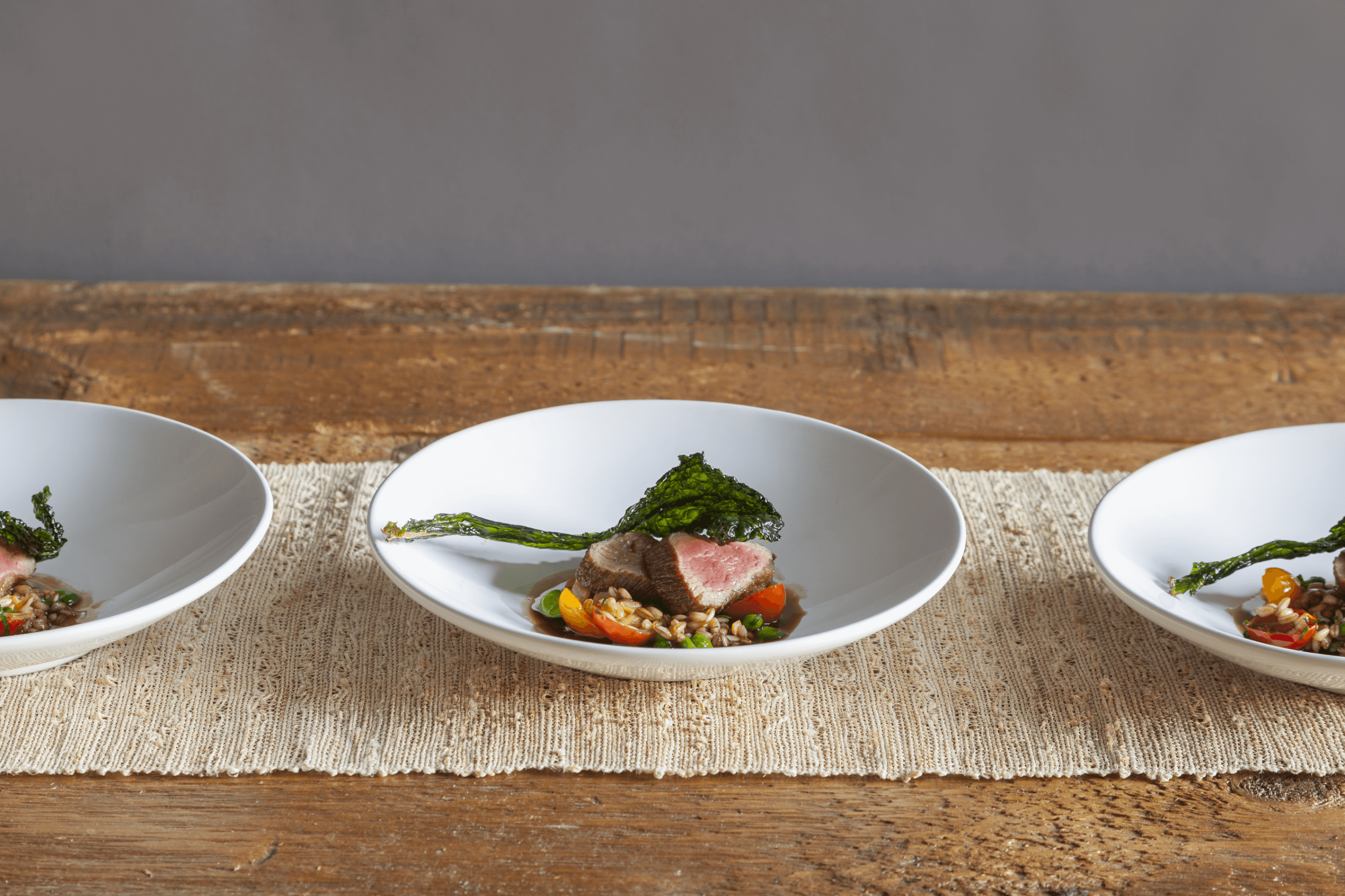 Three plates of gourmet food arranged in a row on a wooden table, each containing sliced meat, grilled vegetables, and greens, presented on a woven table runner.