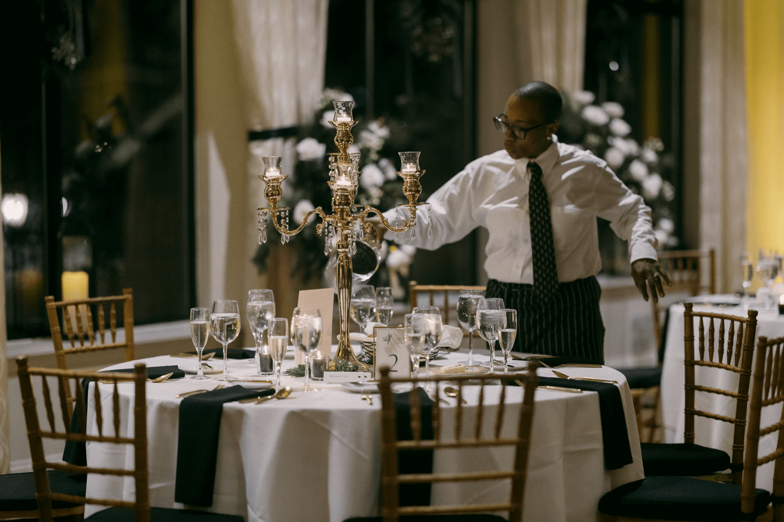 Server arranging a candelabra on a round table set with glasses, napkins, and plates in an elegantly decorated dining room.
