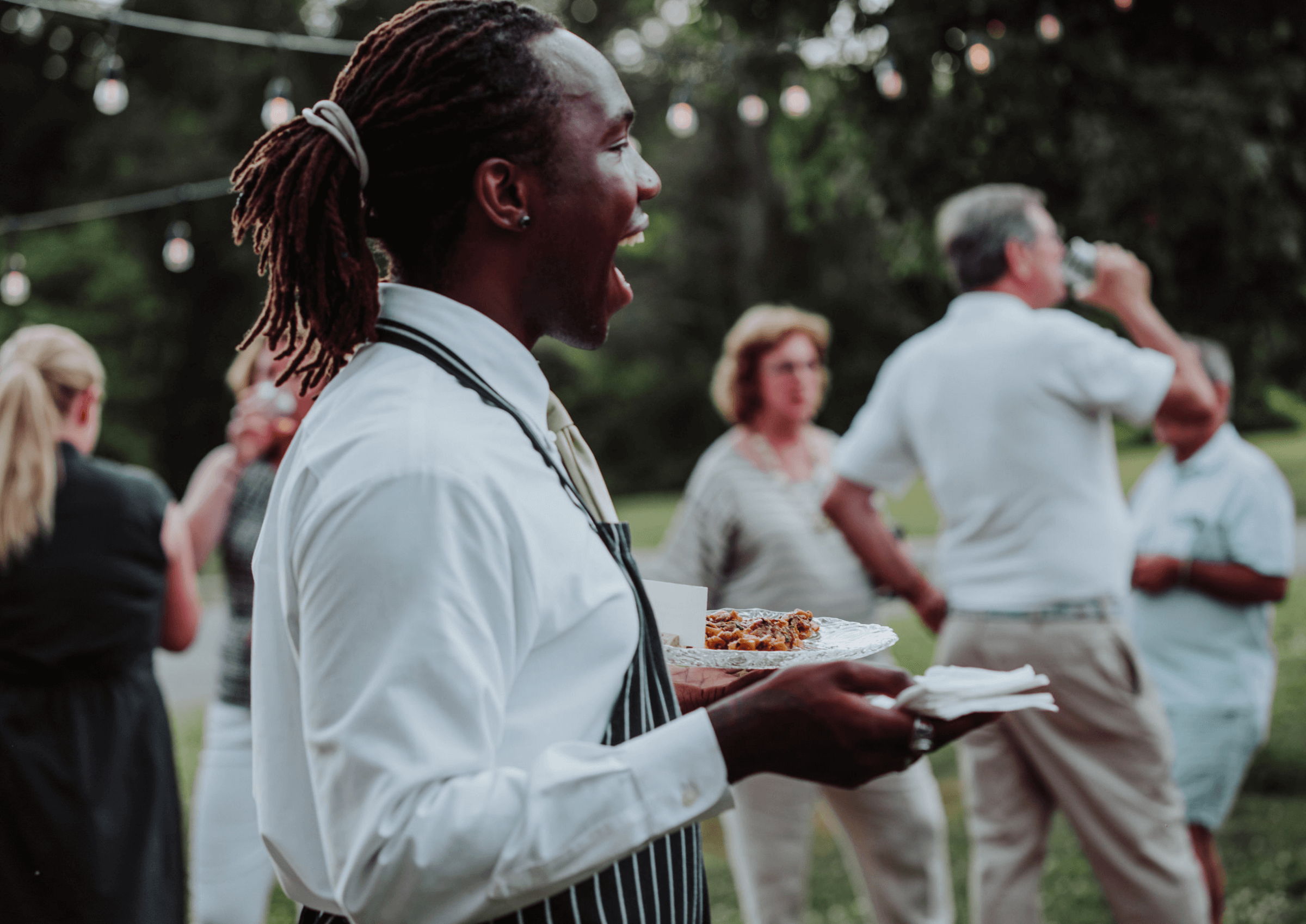 A person in a white shirt and apron stands outdoors at an event, holding a plate of food. Other people are gathered in the background, with string lights overhead.