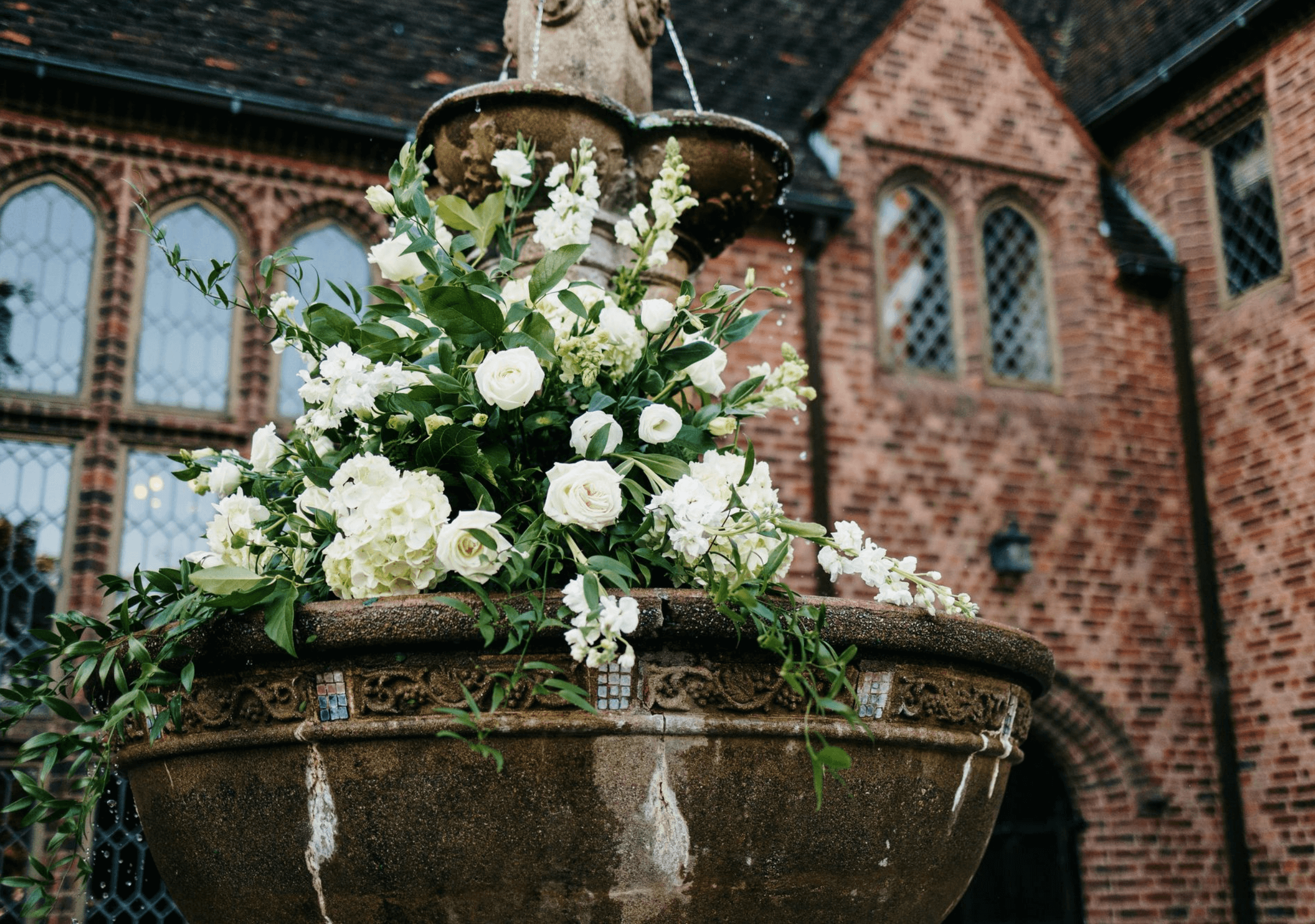 A stone fountain adorned with white flowers and green foliage in front of an old brick building with arched windows.