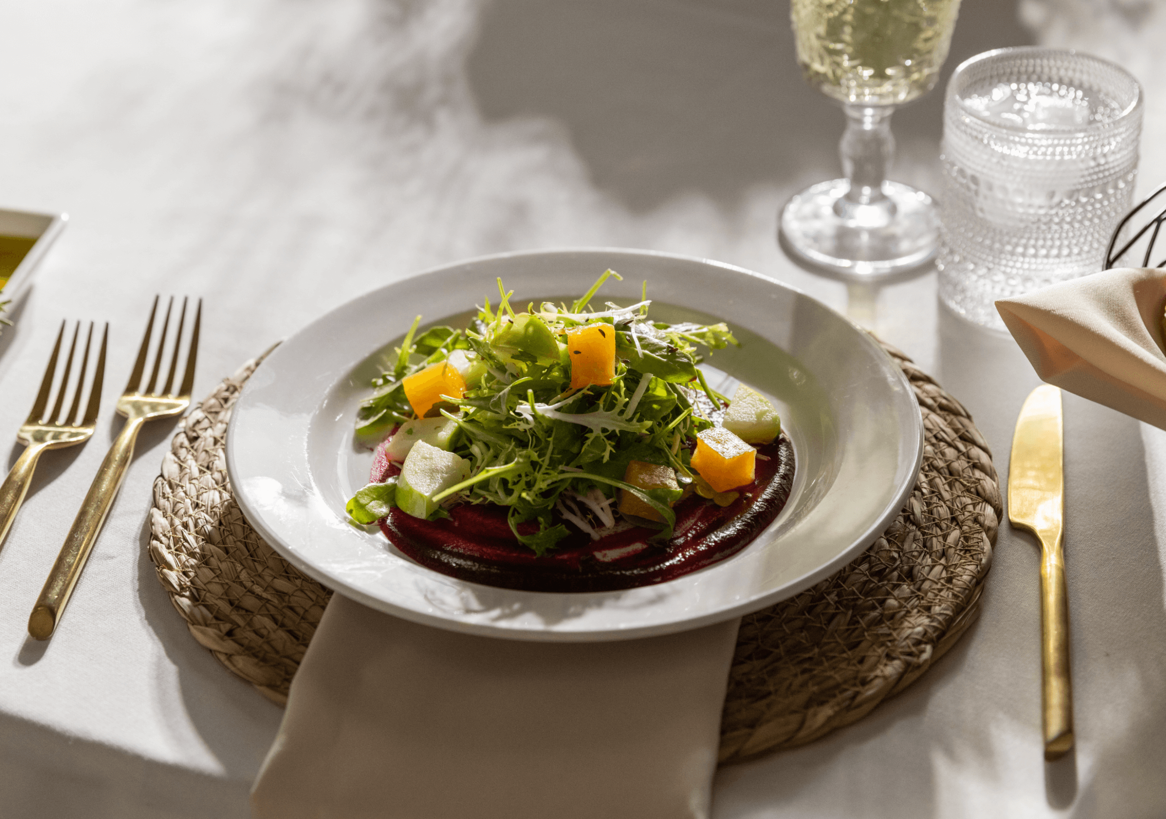 Plate of salad with arugula, beet slices, and orange segments on a woven placemat, surrounded by gold cutlery and a glass of drink in the background.