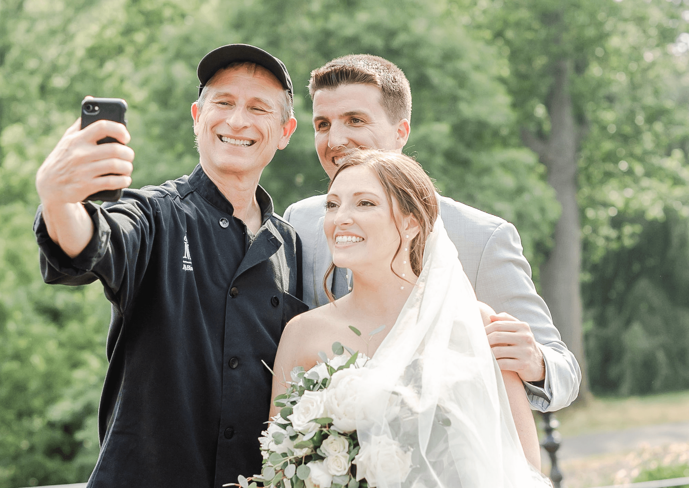 A man in a black chef's coat takes a selfie with a bride holding a bouquet and a groom in a suit, all smiling. Trees are visible in the background.