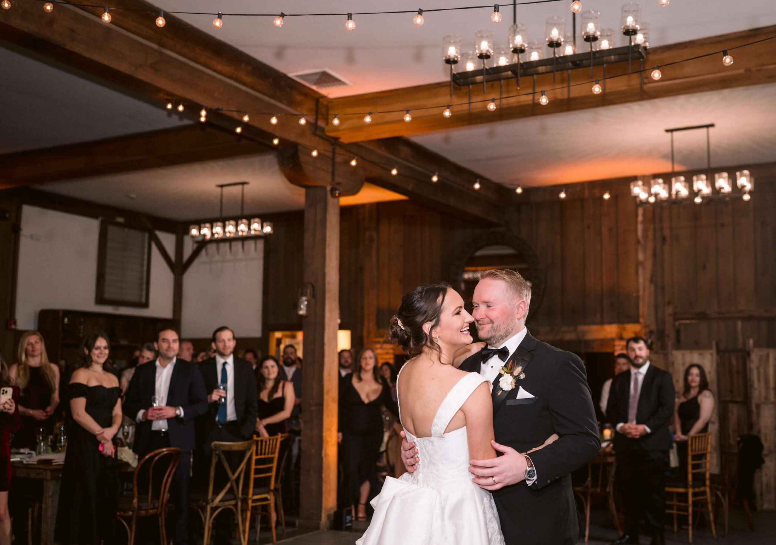 A bride and groom share their first dance in a warmly lit venue, surrounded by seated and standing guests.