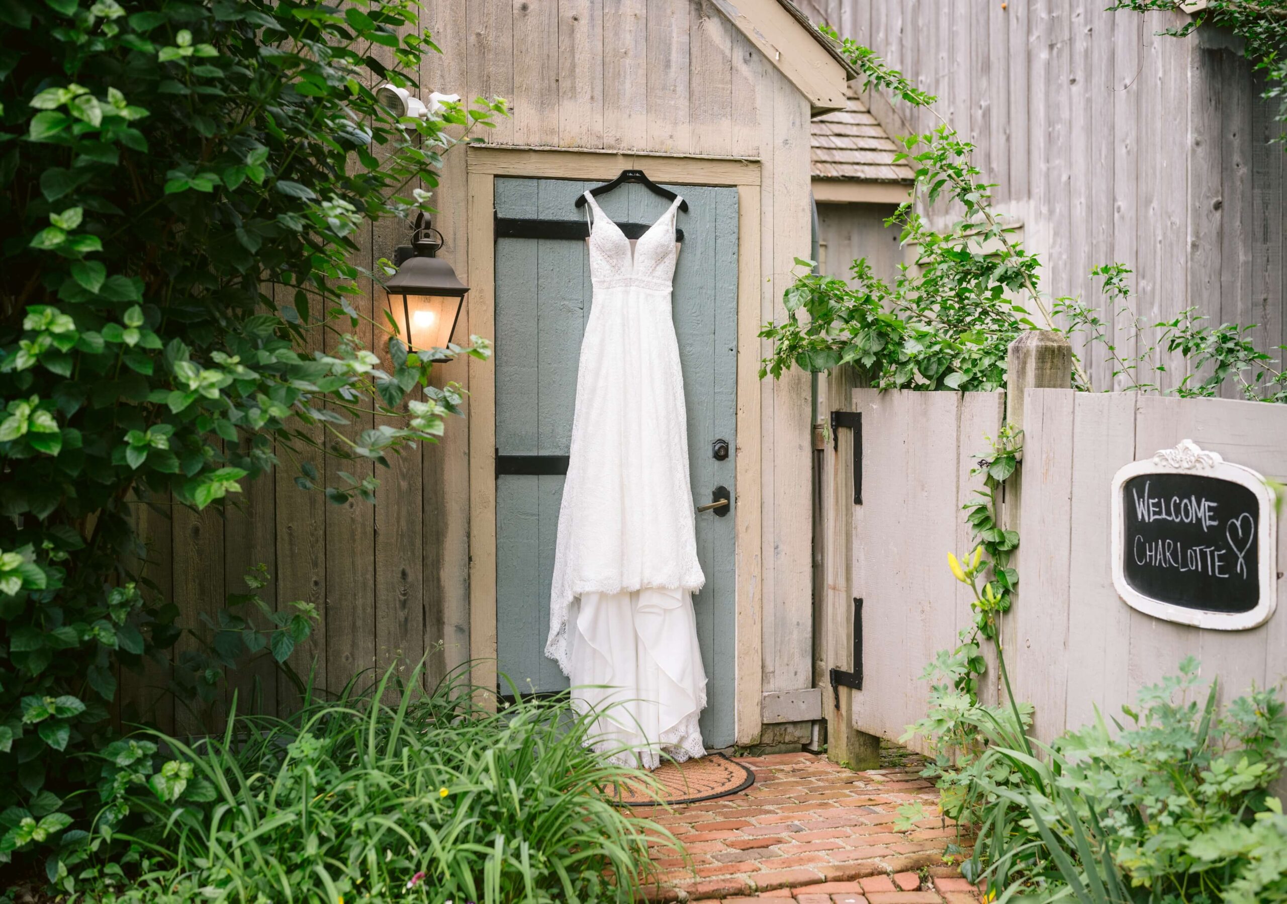 White lace wedding dress hanging on a wooden door outside, surrounded by greenery and a chalkboard sign reading 