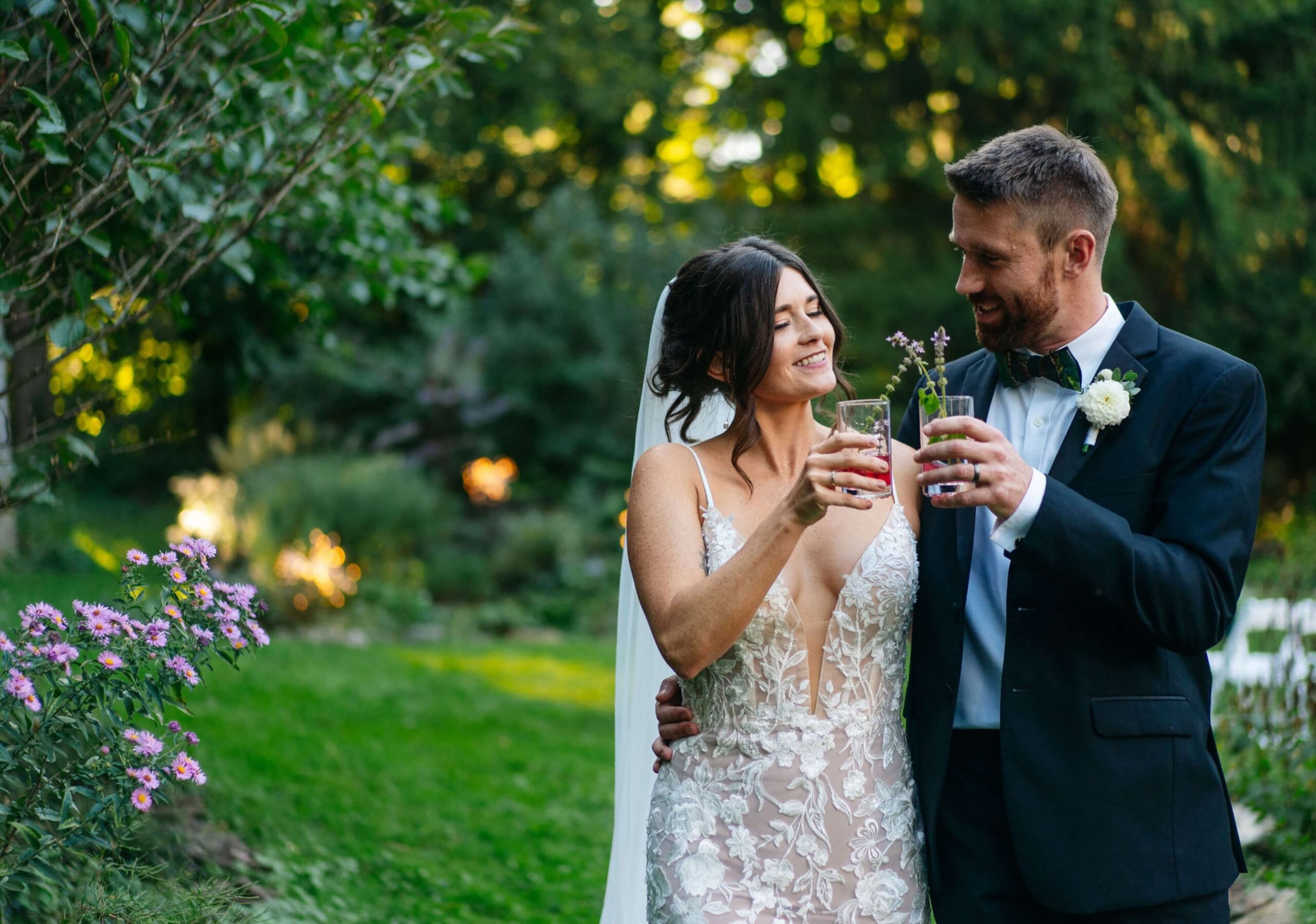 Bride and groom in formal attire toast with drinks in a lush garden setting.