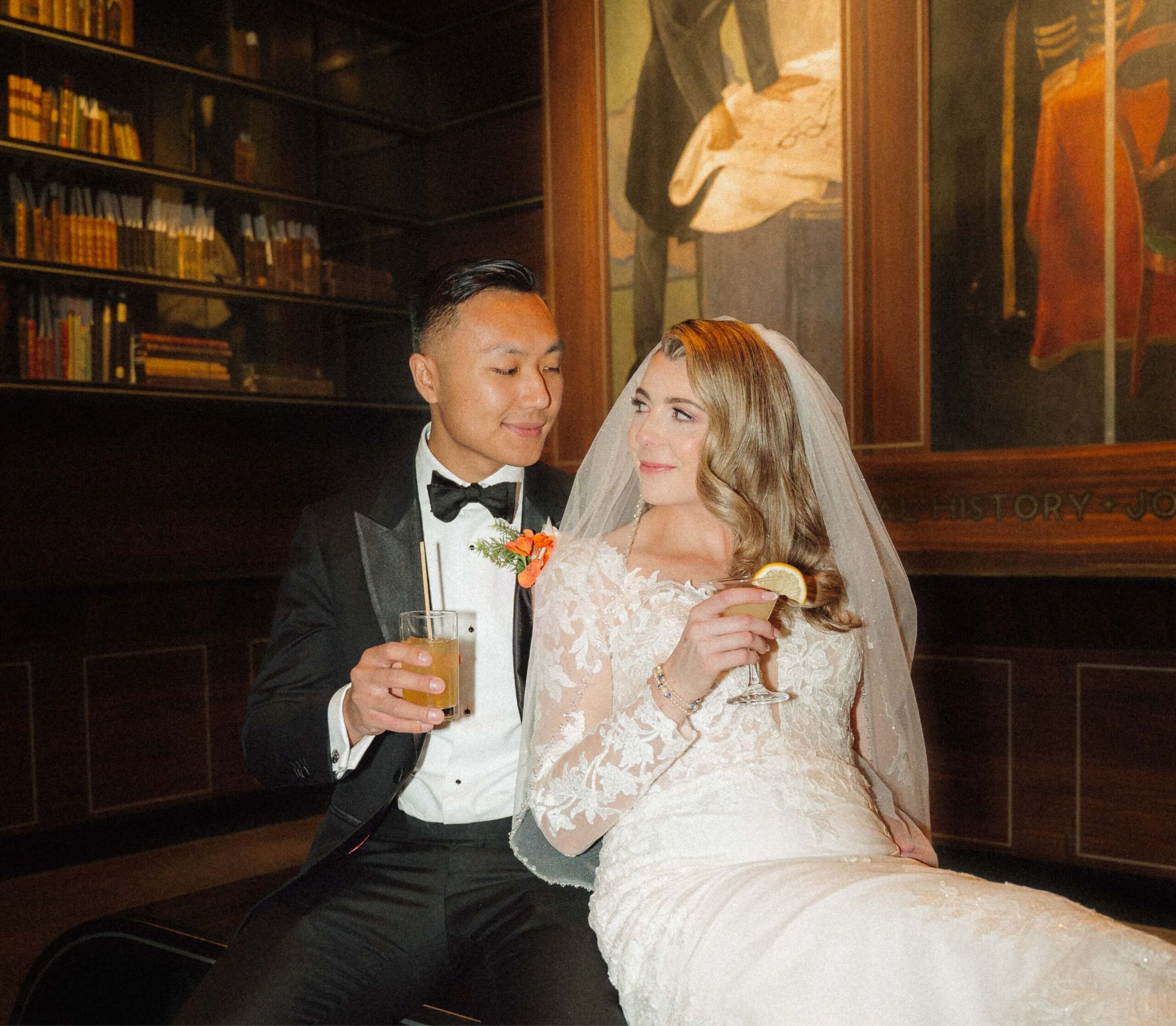 A bride and groom in formal attire sit together, holding drinks, in a room lined with books and paintings.