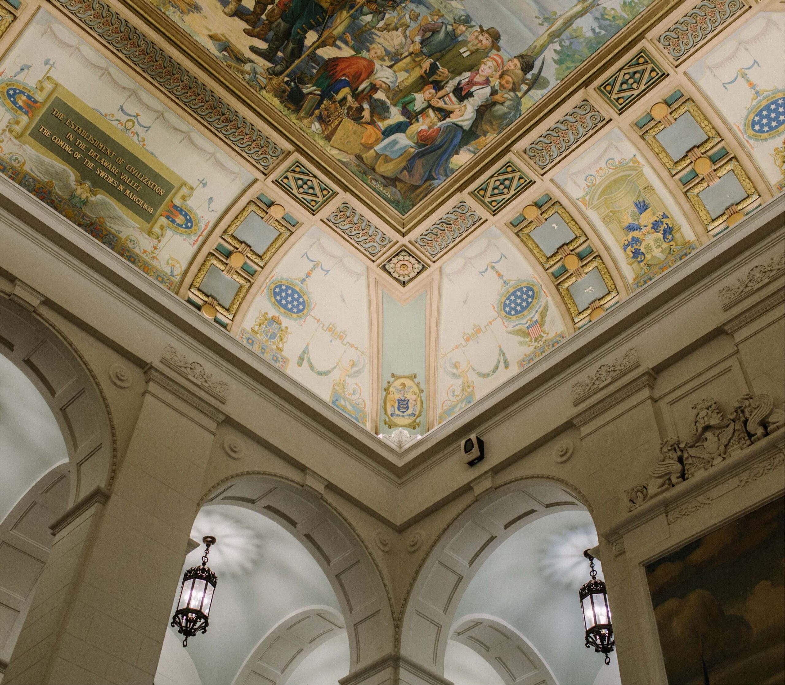 Ornate architectural ceiling with colorful murals and chandeliers in a grand hall. Arched doorways and decorative moldings are visible.