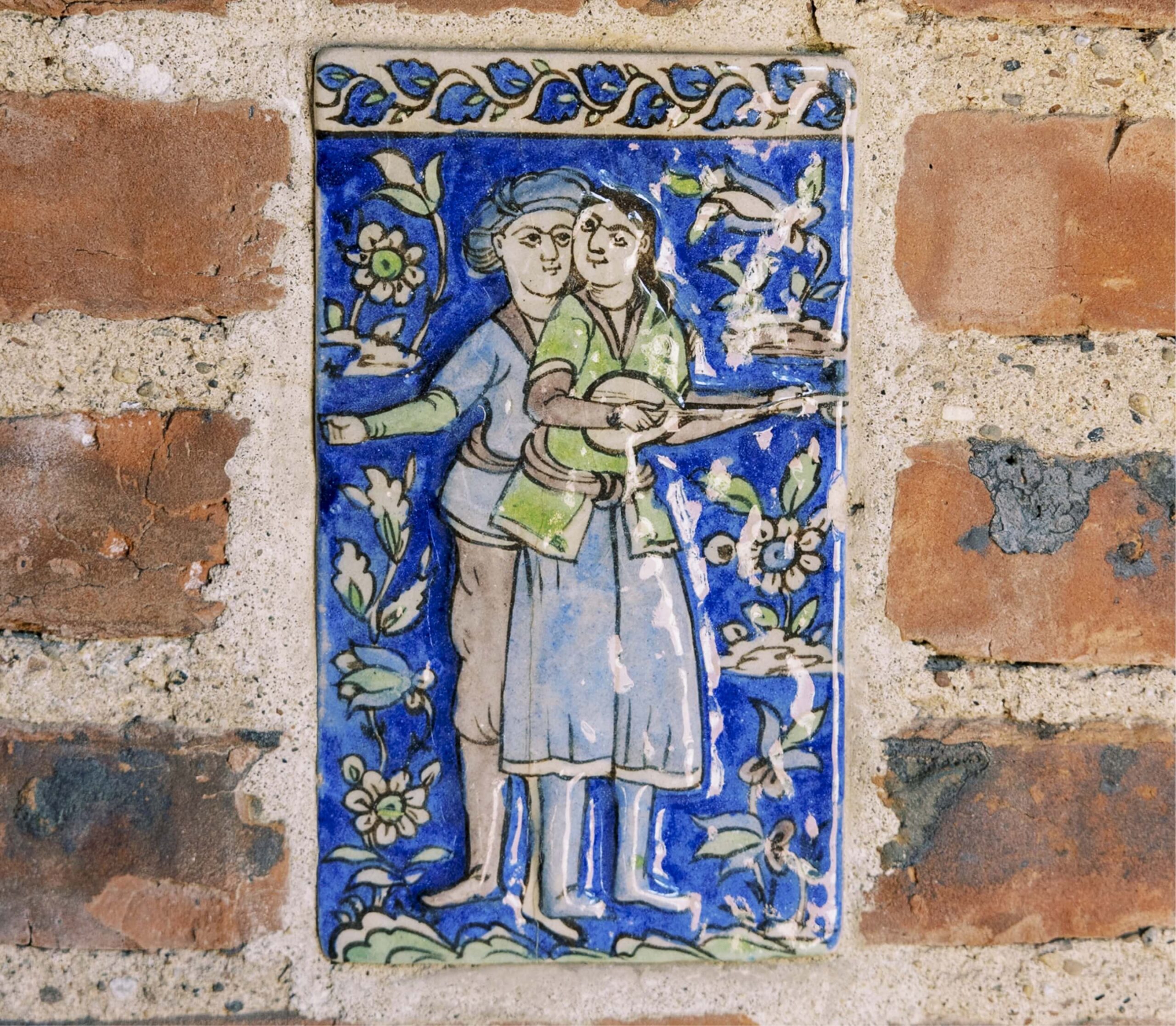 Ceramic tile on brick wall depicting two figures, one holding a string instrument, surrounded by floral patterns on a blue background.