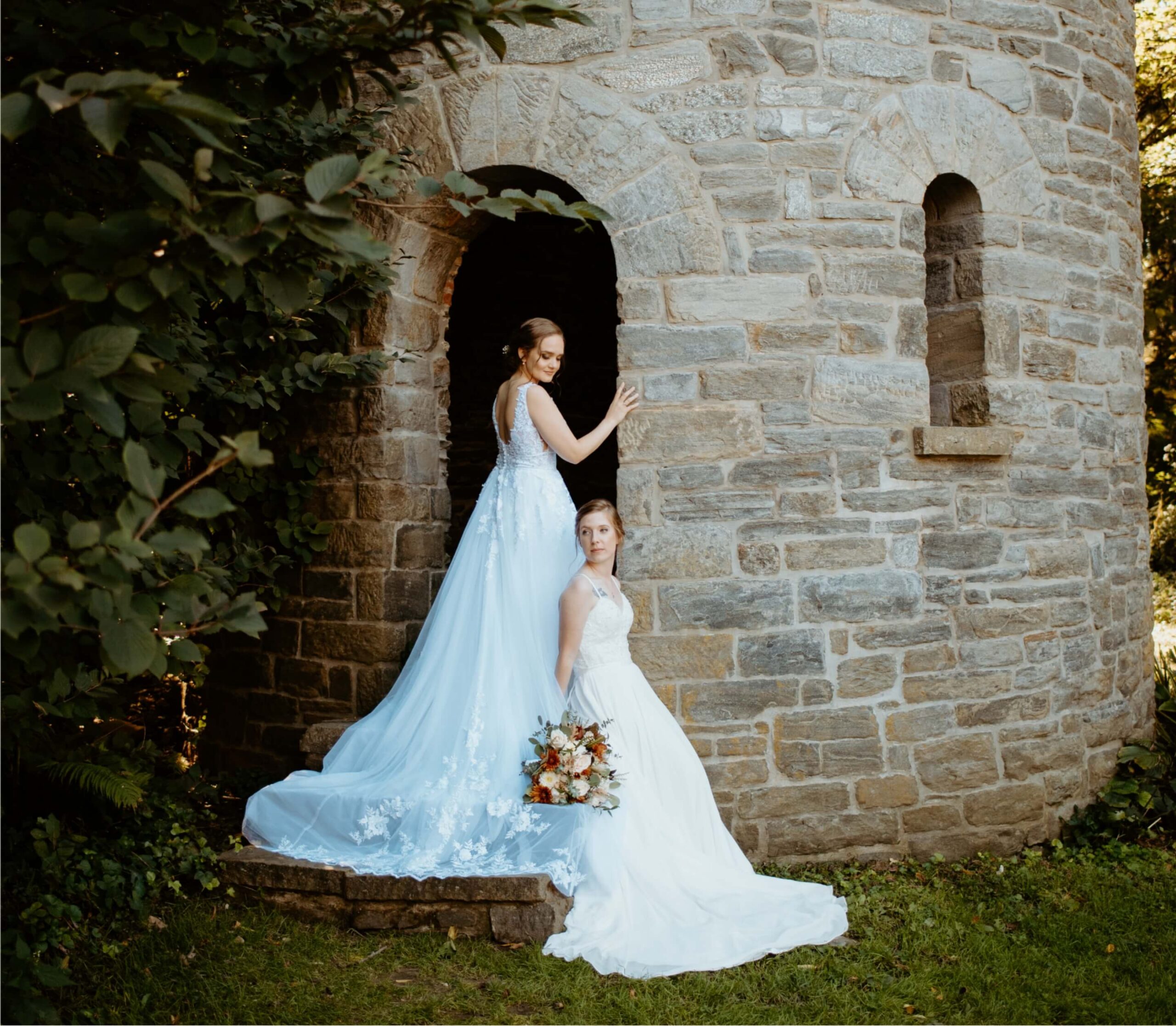 Two people in white gowns stand by a stone tower, one seated holding a bouquet. The background features greenery and sunlight.