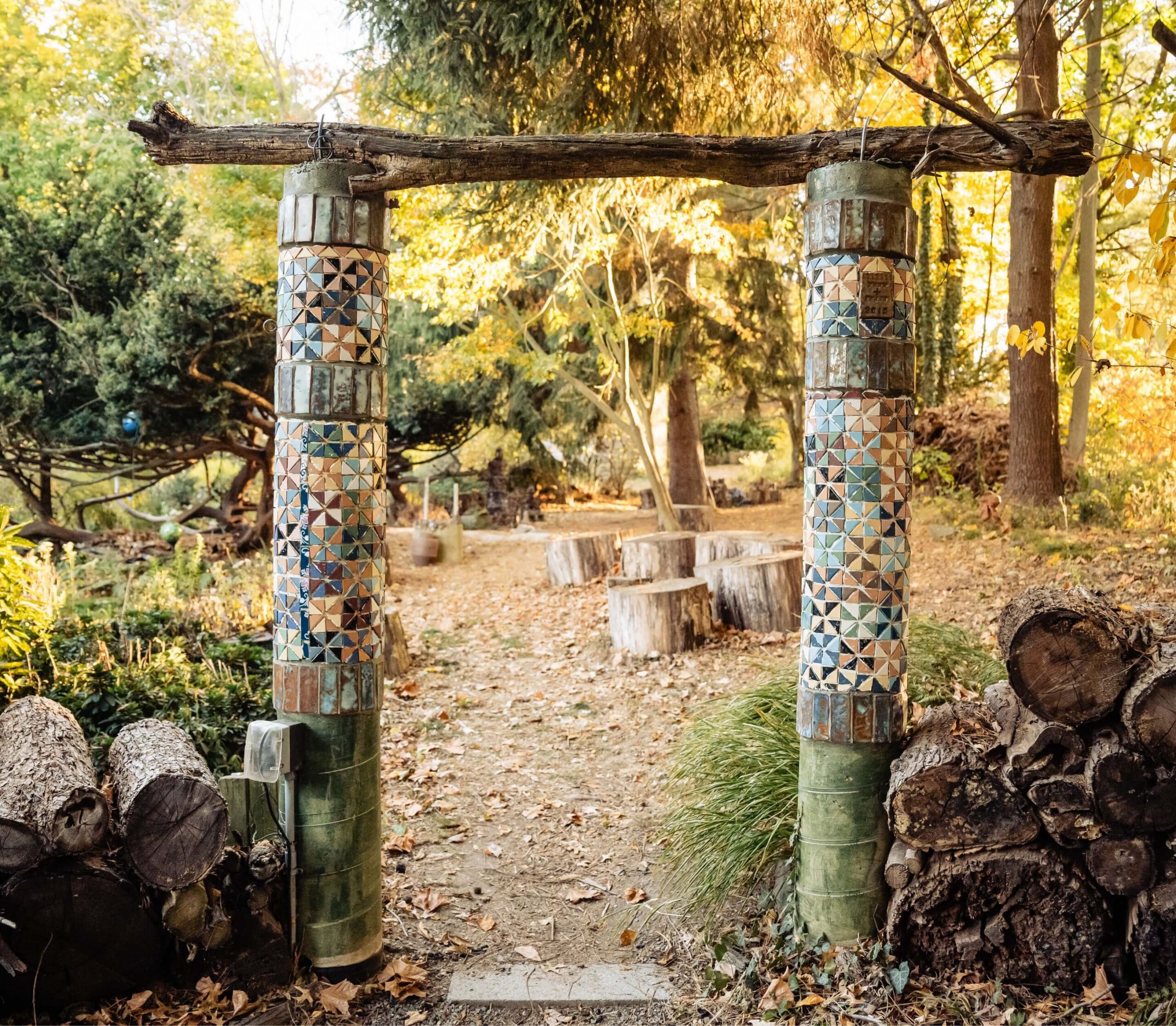 Mosaic pillar gate made of colorful tiles and driftwood with a dirt path leading through a forested area surrounded by logs and trees.