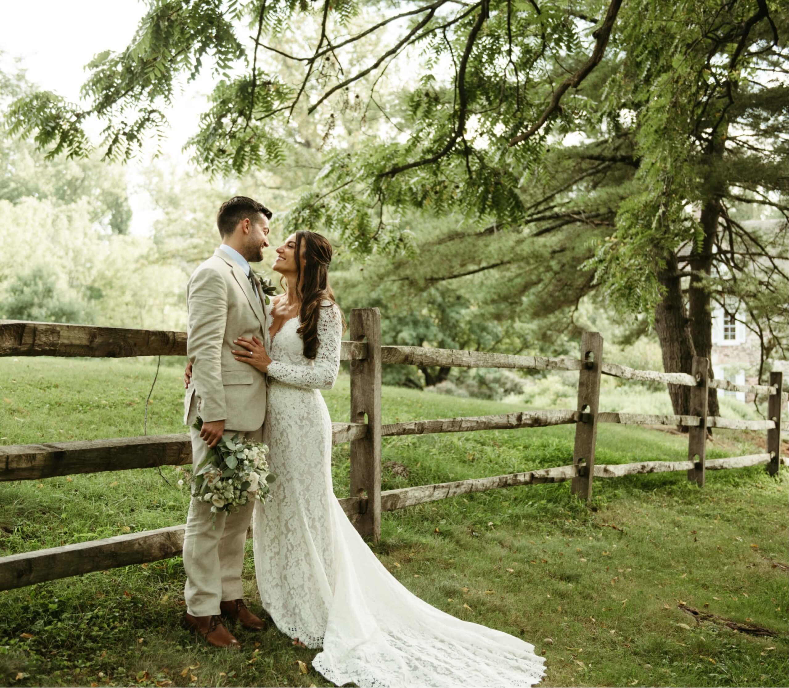 A bride in a lace gown and a groom in a beige suit stand together by a wooden fence in a grassy area with trees. The bride holds a bouquet.