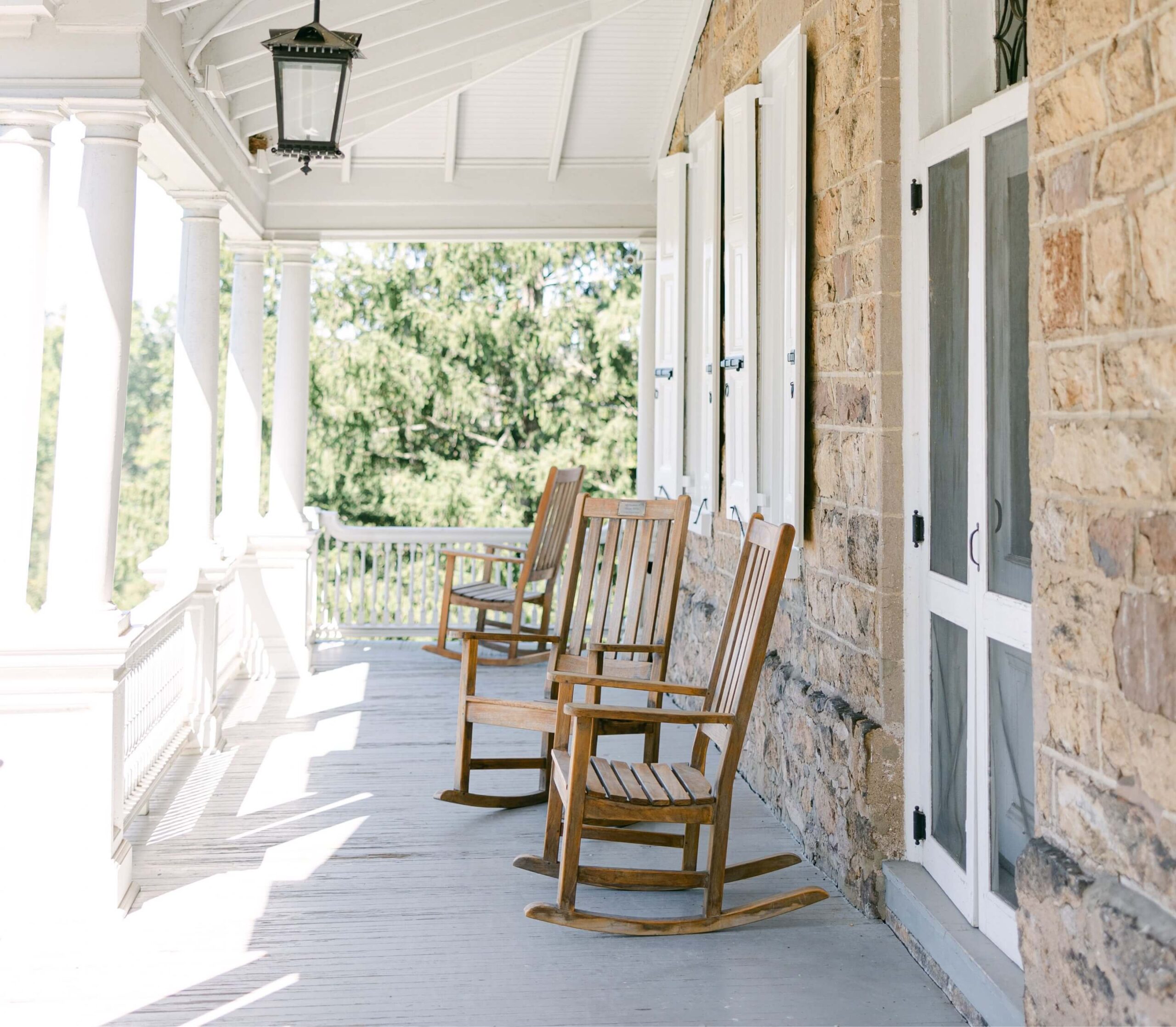 A shaded porch with three wooden rocking chairs and a stone wall background, overlooking a green landscape. A lantern hangs from the white ceiling.