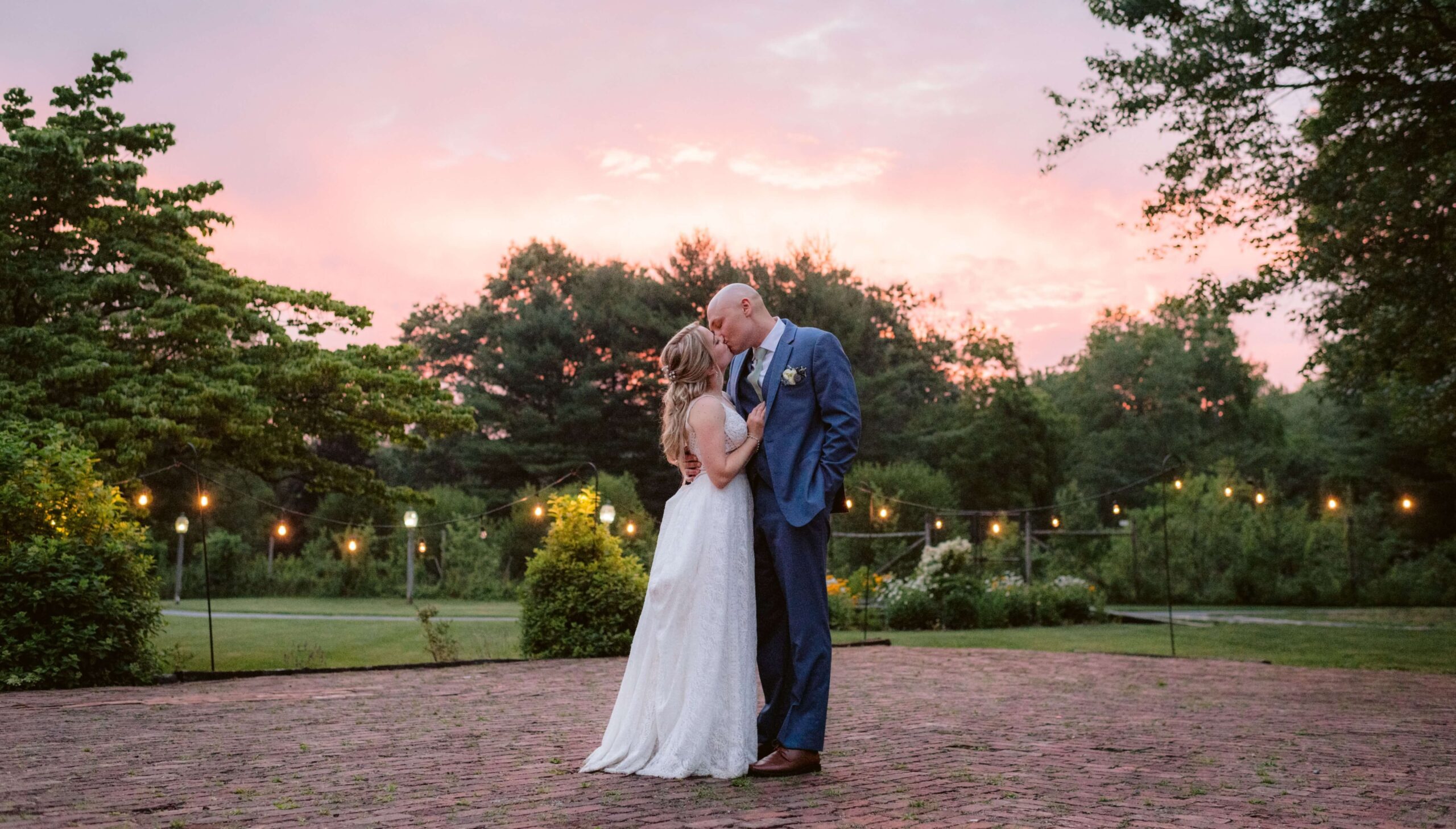 A couple in formal attire embraces on a brick pavement at sunset. String lights and trees are visible in the background.