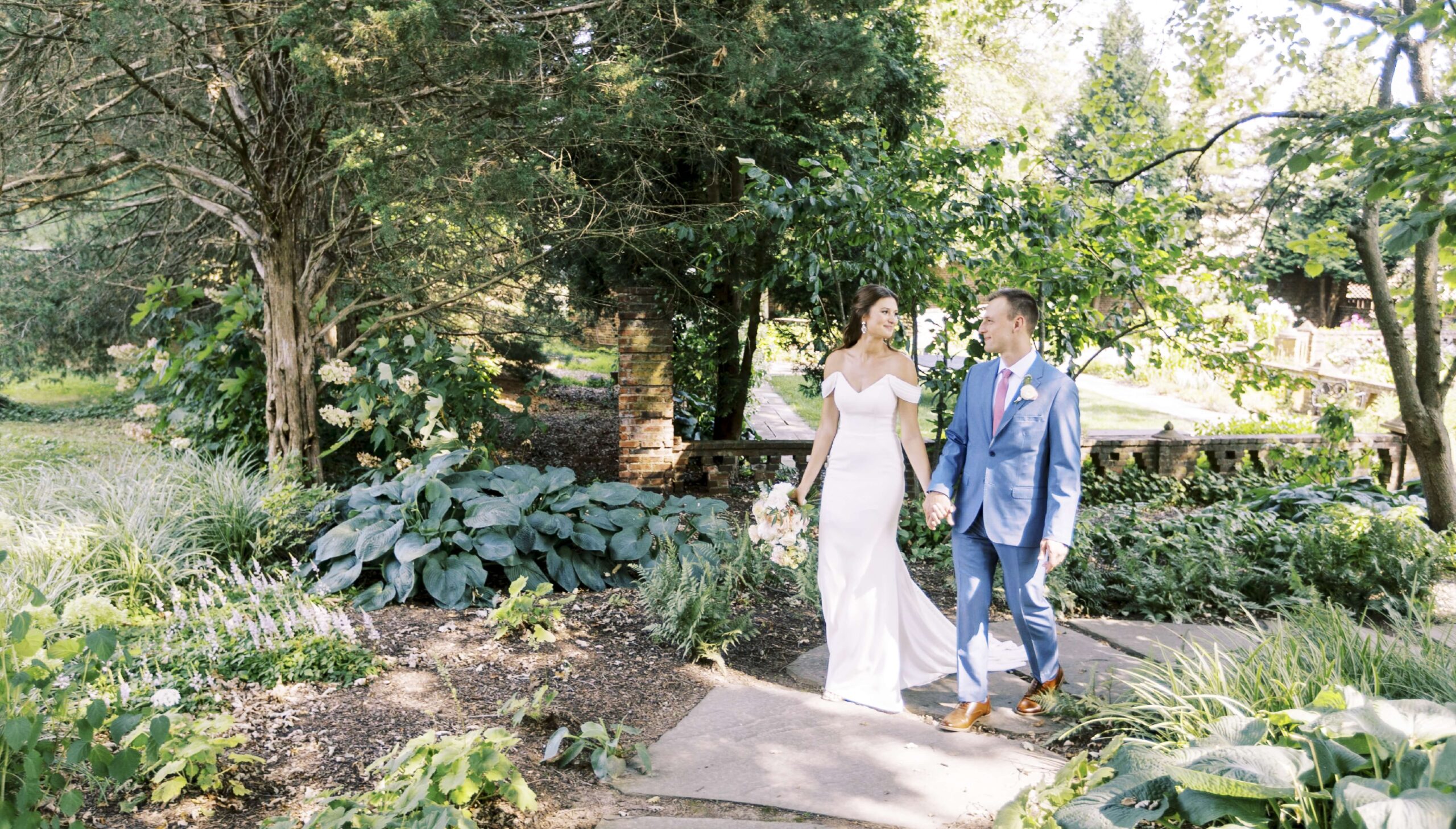 A bride in a white dress and a groom in a blue suit holding hands, walking along a path surrounded by greenery.
