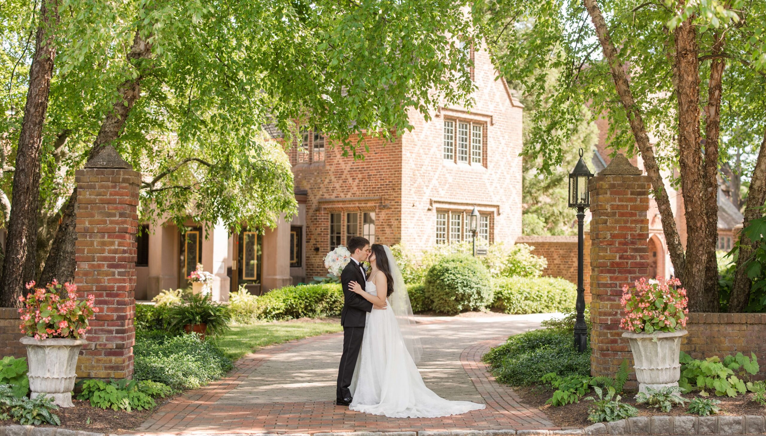 A bride and groom embrace on a brick path in front of a large, lush garden and a brick house surrounded by trees.