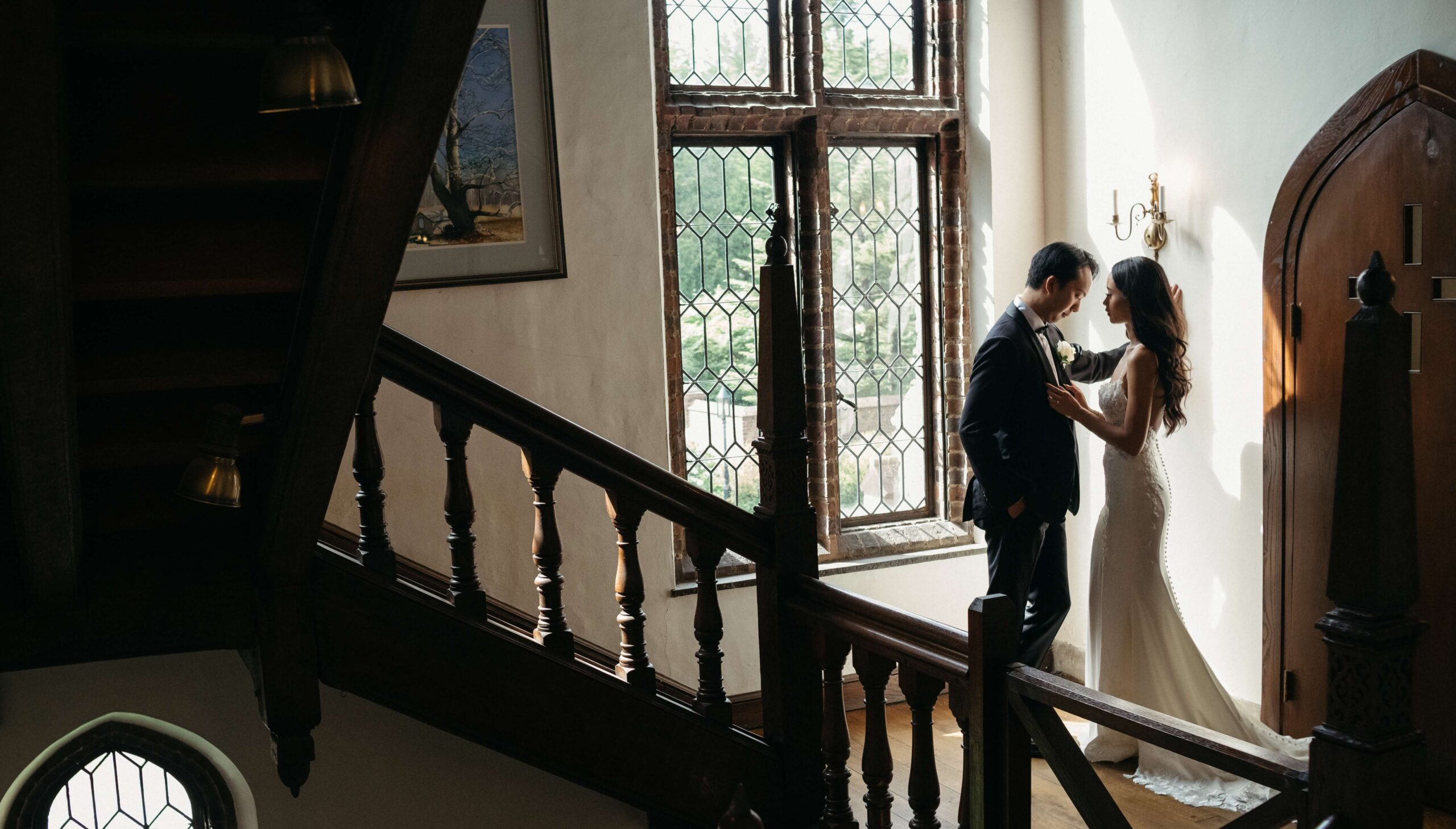 A couple stands by a large window in a dimly lit room with wooden stairs and a chandelier, gazing at each other.