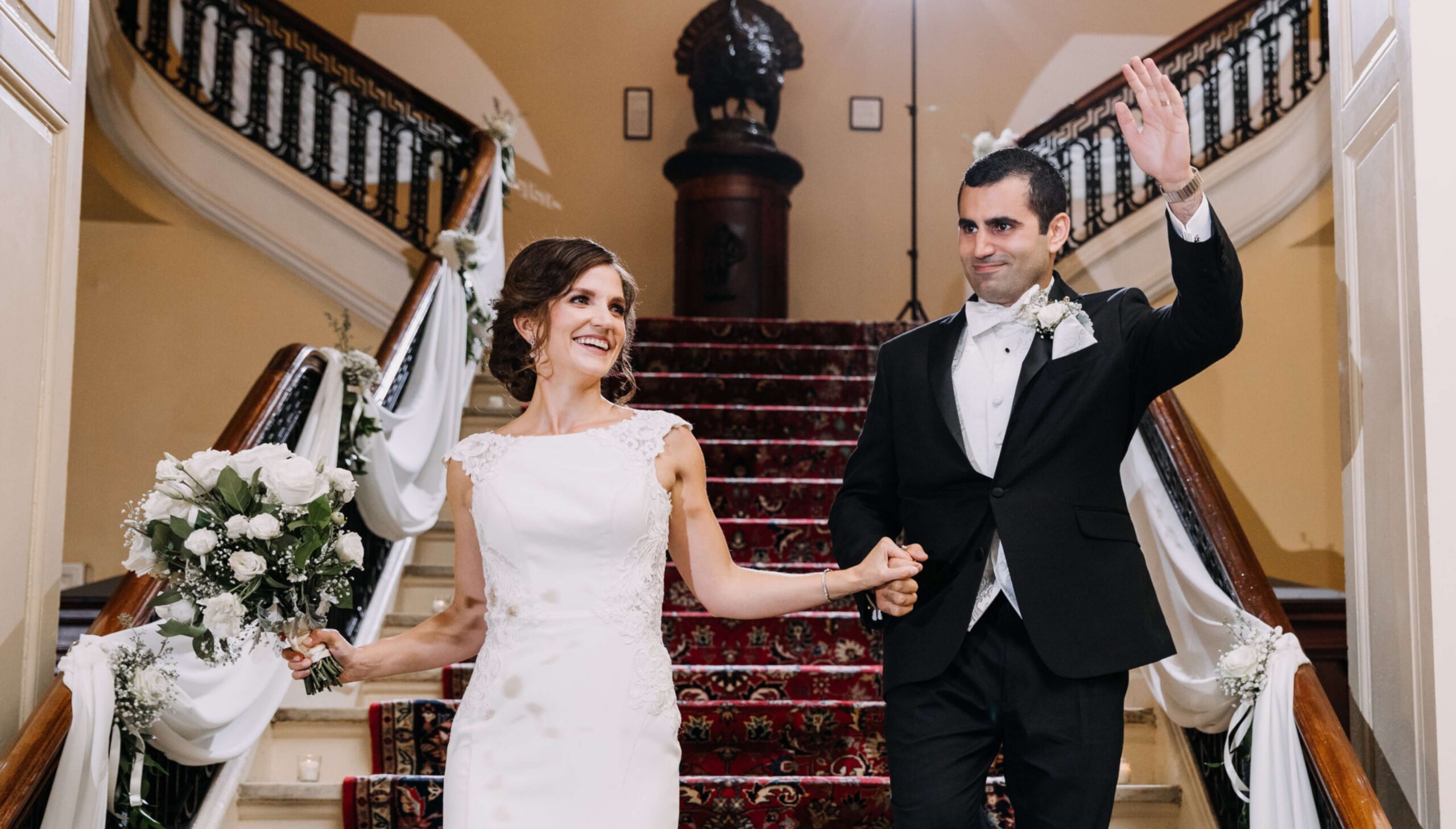 A bride and groom hold hands and wave as they descend a staircase decorated with white flowers and fabric.