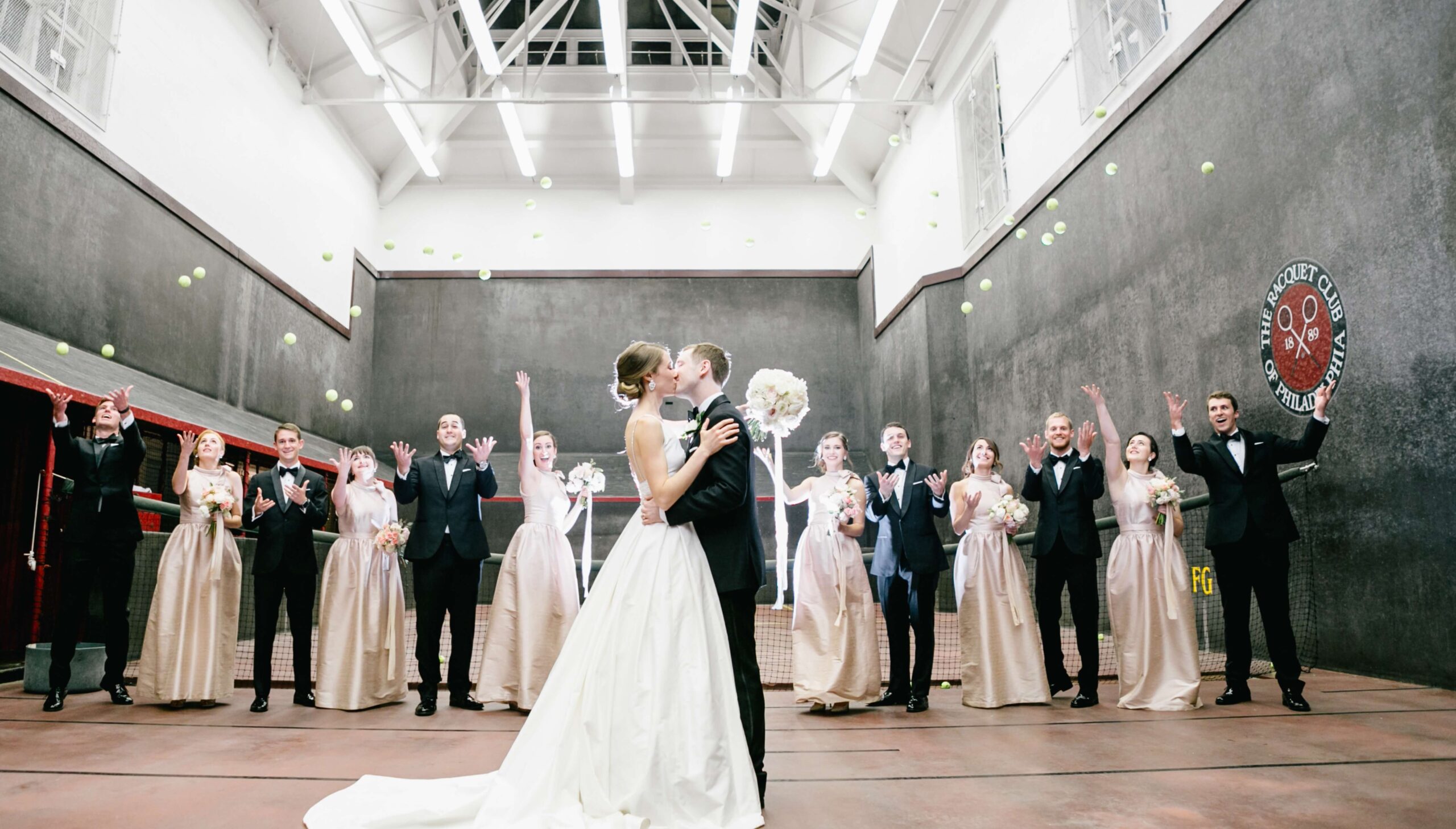 A bride and groom kiss on an indoor tennis court while bridesmaids and groomsmen throw tennis balls in the air.