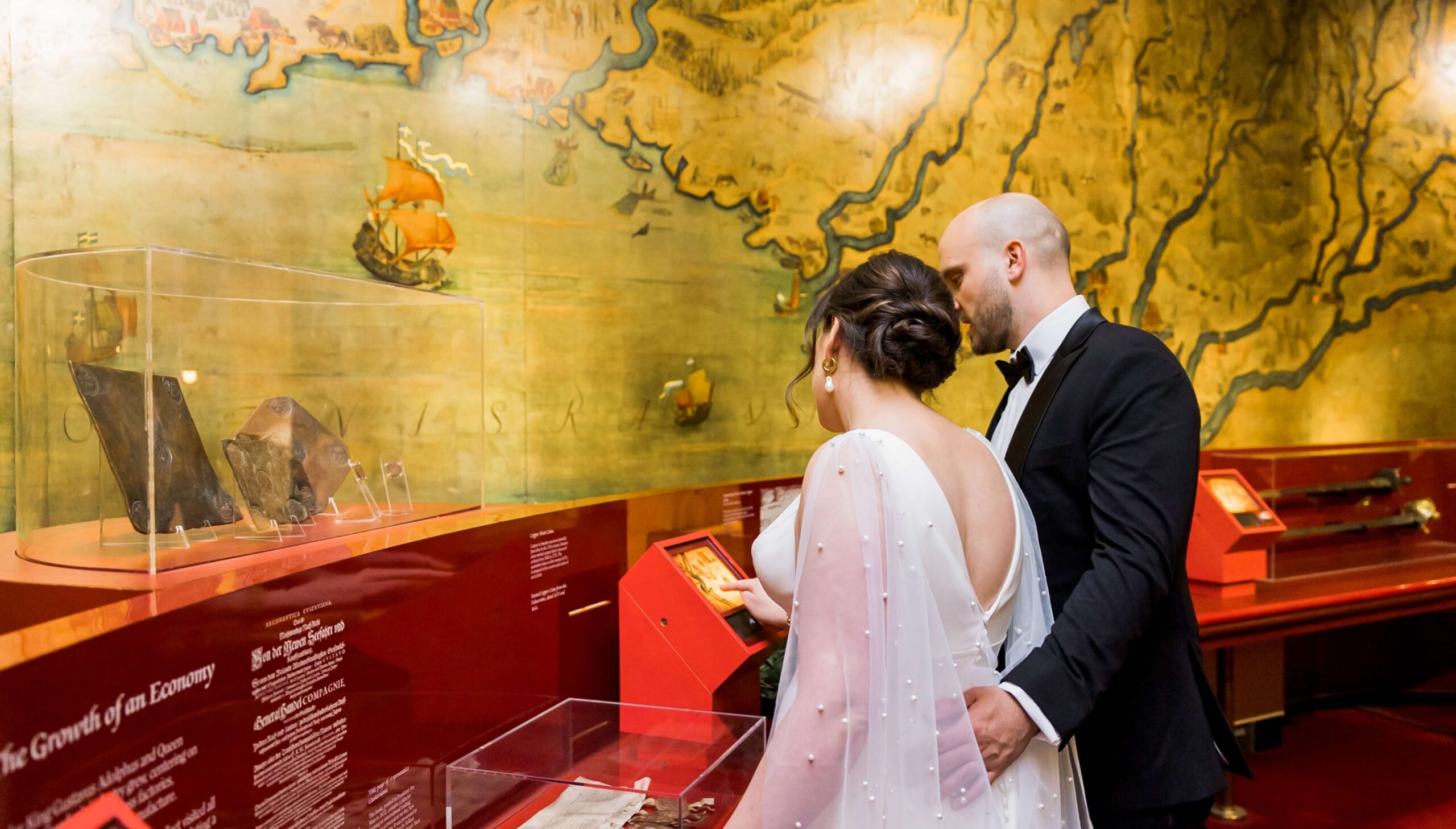 A couple in formal attire views displays in a museum. The background features a large, historical map and a ship mural.
