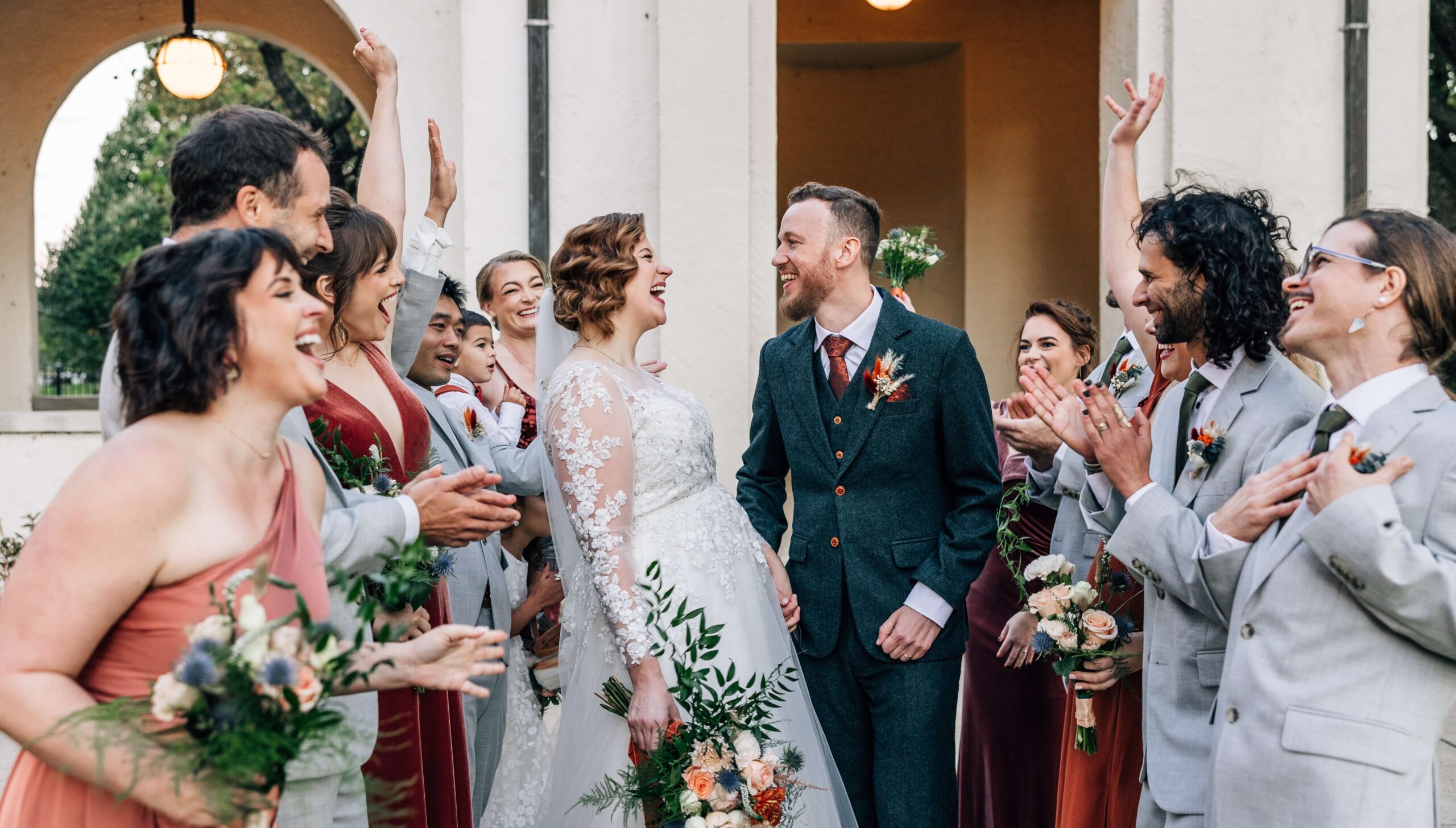 A bride and groom smile at each other, surrounded by a joyful wedding party in colorful attire, holding flowers, in front of a building.