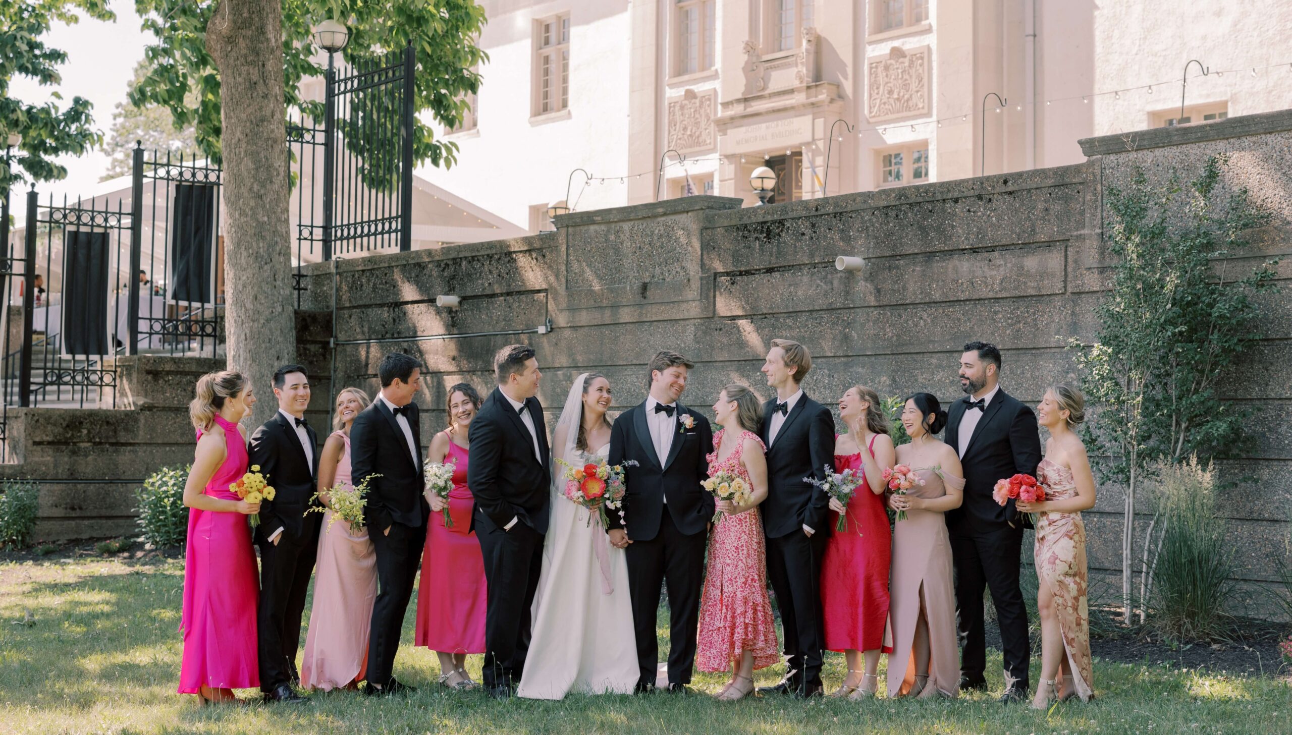 A wedding party stands outdoors in formal attire. The group includes bridesmaids in pink and red dresses, groomsmen in black suits, and a bride and groom in the center holding flowers.