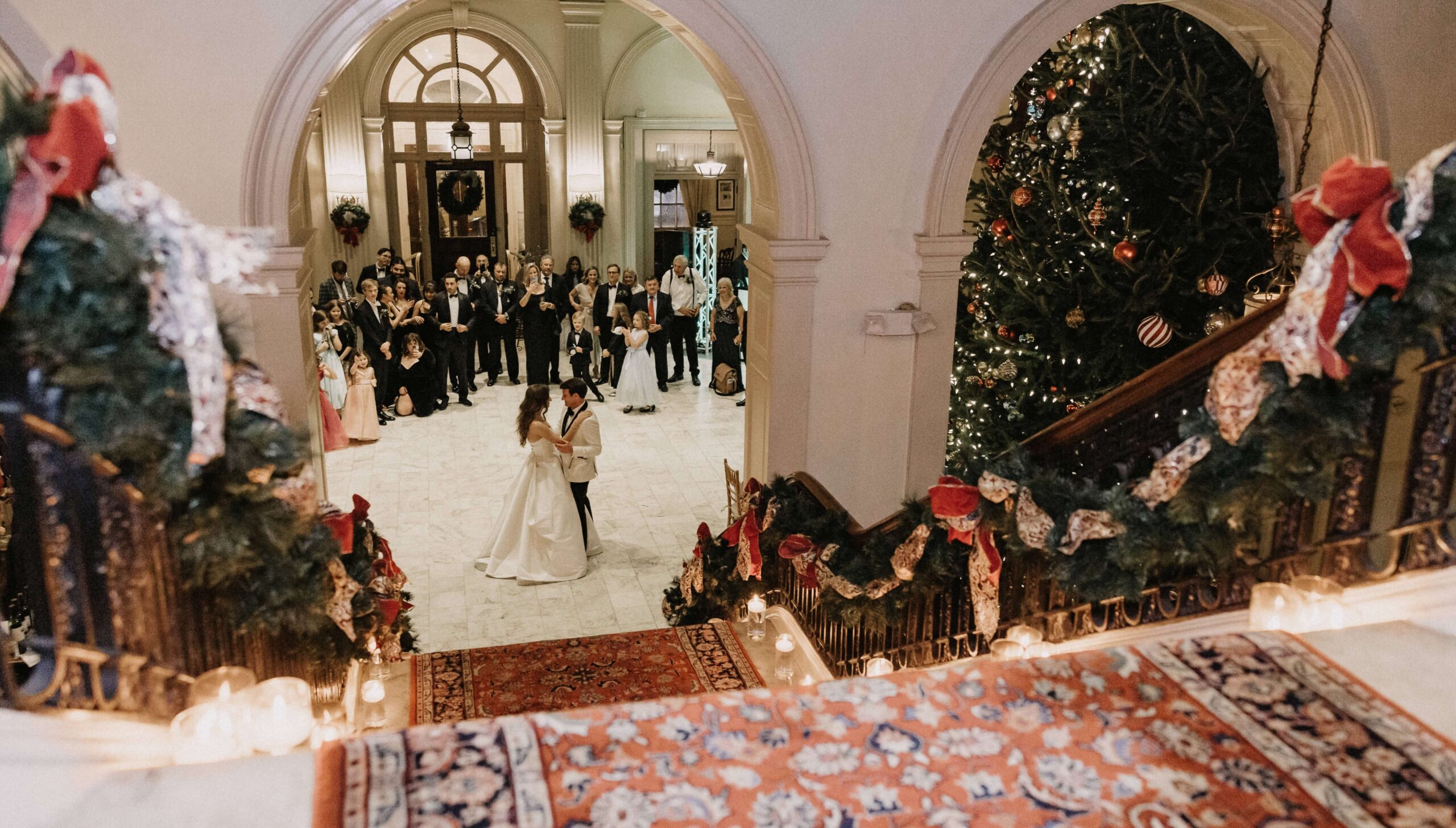 A couple dances at the foot of a grand staircase during a wedding ceremony. Guests watch from a distance, and a decorated Christmas tree is visible nearby.