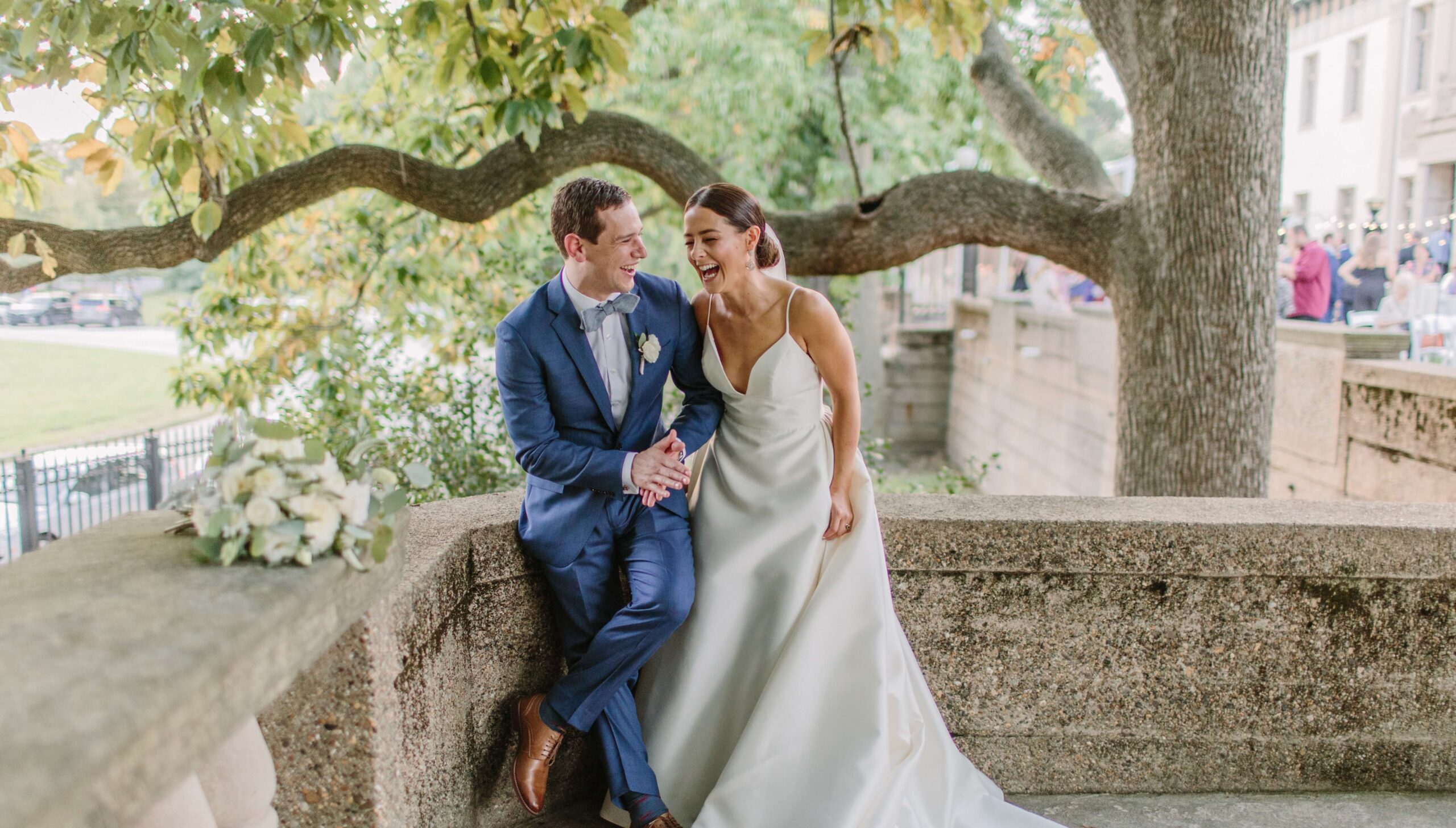 A bride and groom sit on a stone ledge outdoors, under a tree, smiling and holding hands. A bouquet rests beside them.