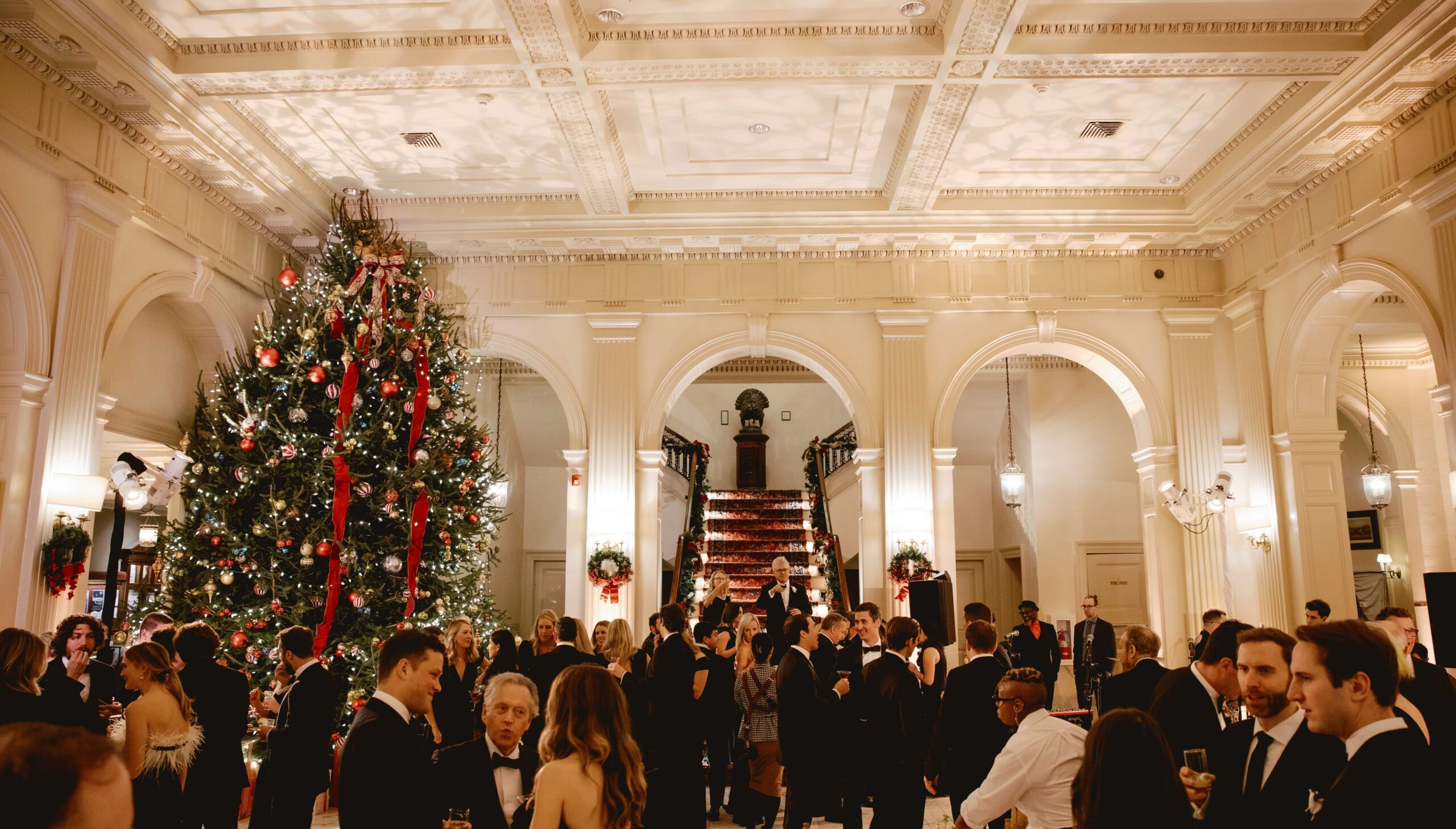 Guests in formal attire mingle at a holiday party in an elegant venue with a large decorated Christmas tree and grand staircase.