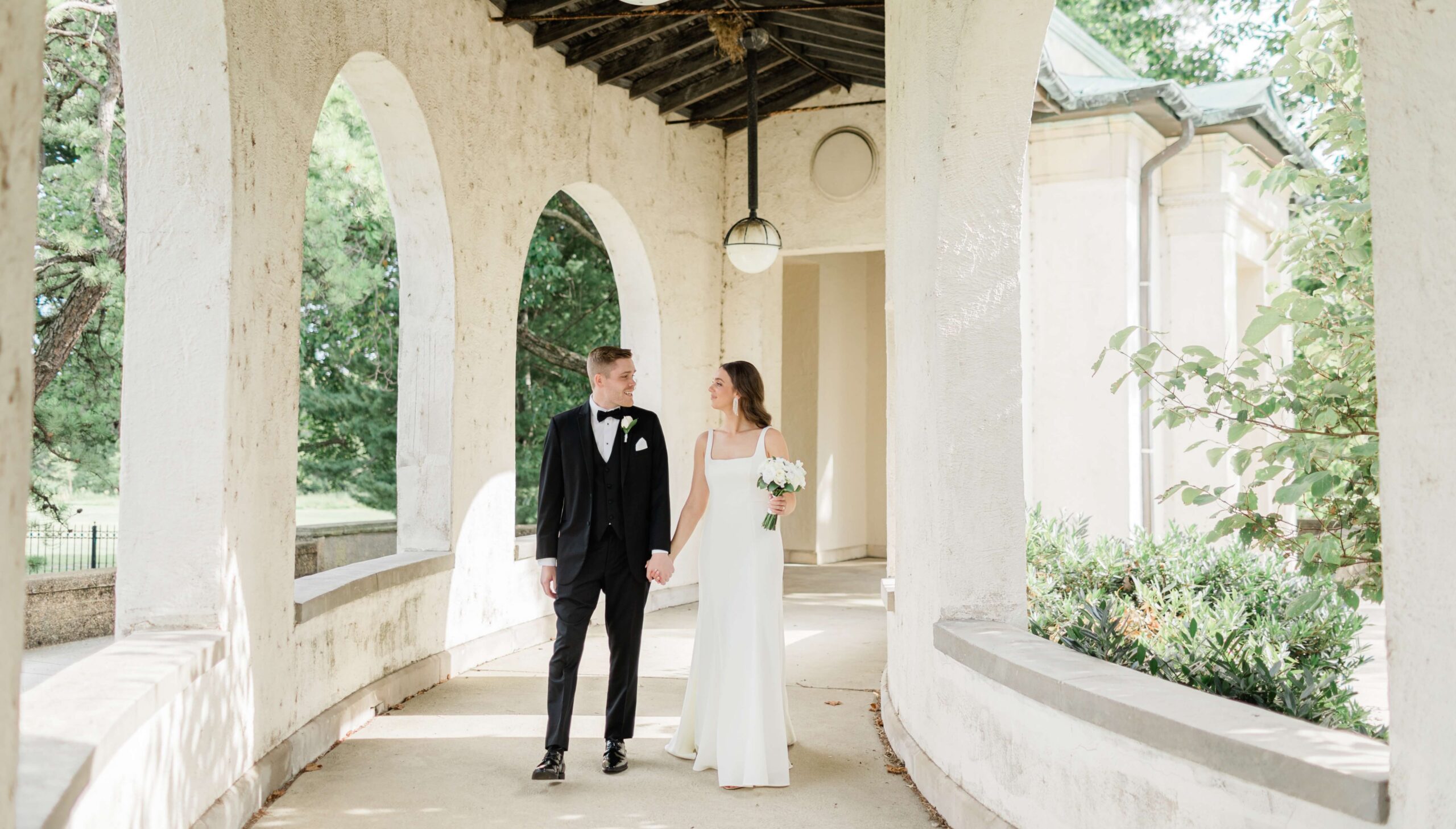 A couple in formal attire holds hands, walking through an arched corridor. The man wears a black suit, and the woman is in a white dress holding a bouquet.
