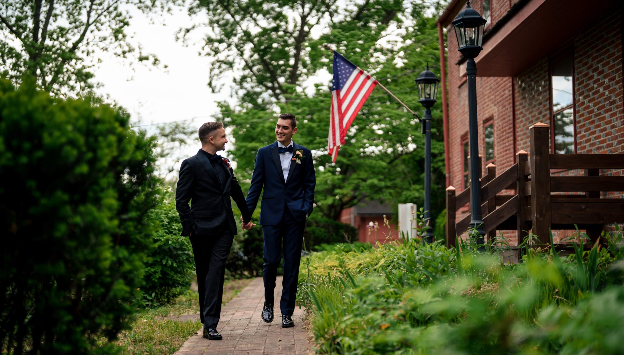 Two people in suits walk hand in hand down a garden path, with an American flag and a brick building in the background.
