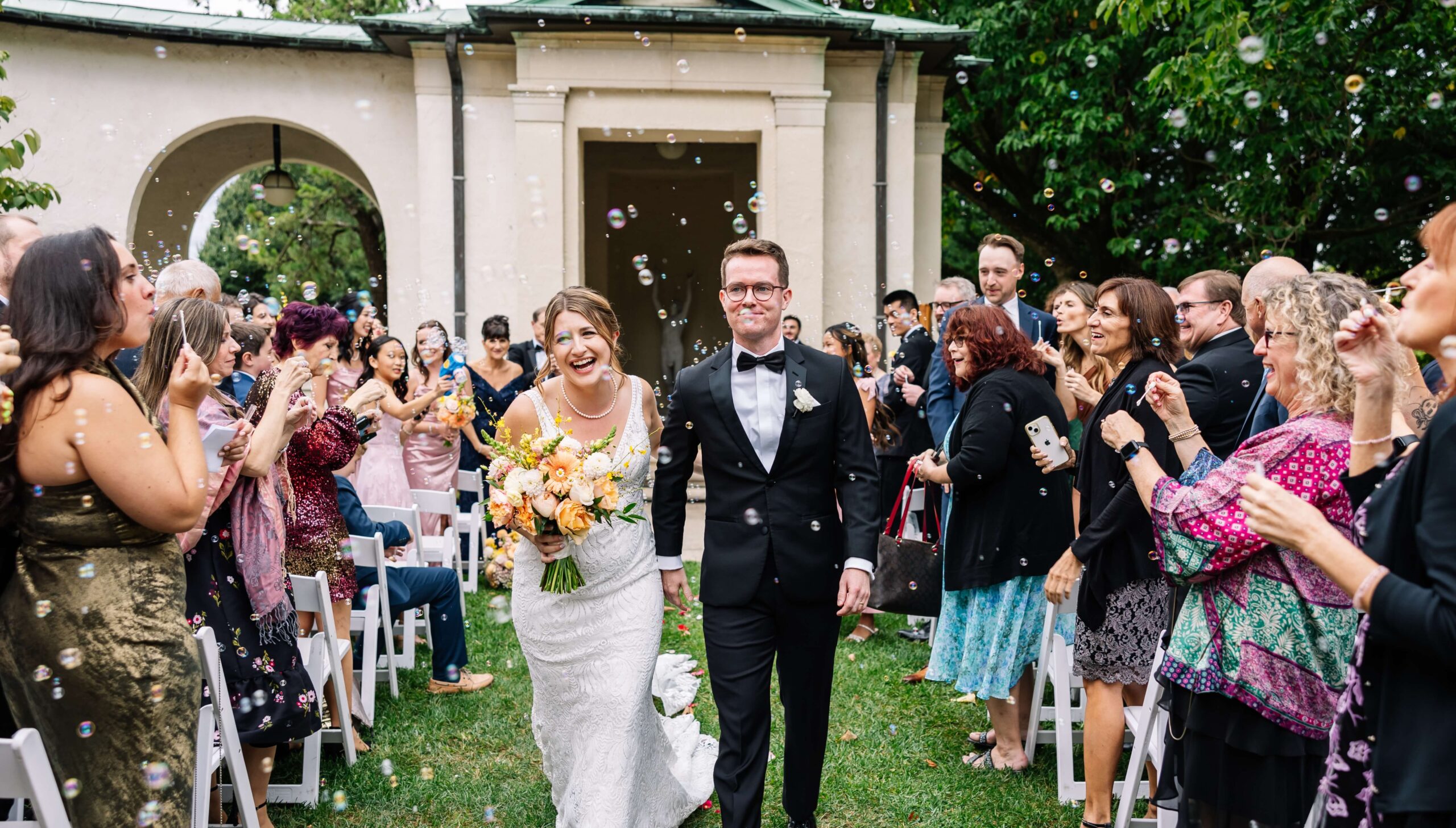 A bride and groom walk down an outdoor aisle, smiling, surrounded by guests blowing bubbles and clapping. The bride holds a bouquet of yellow flowers.
