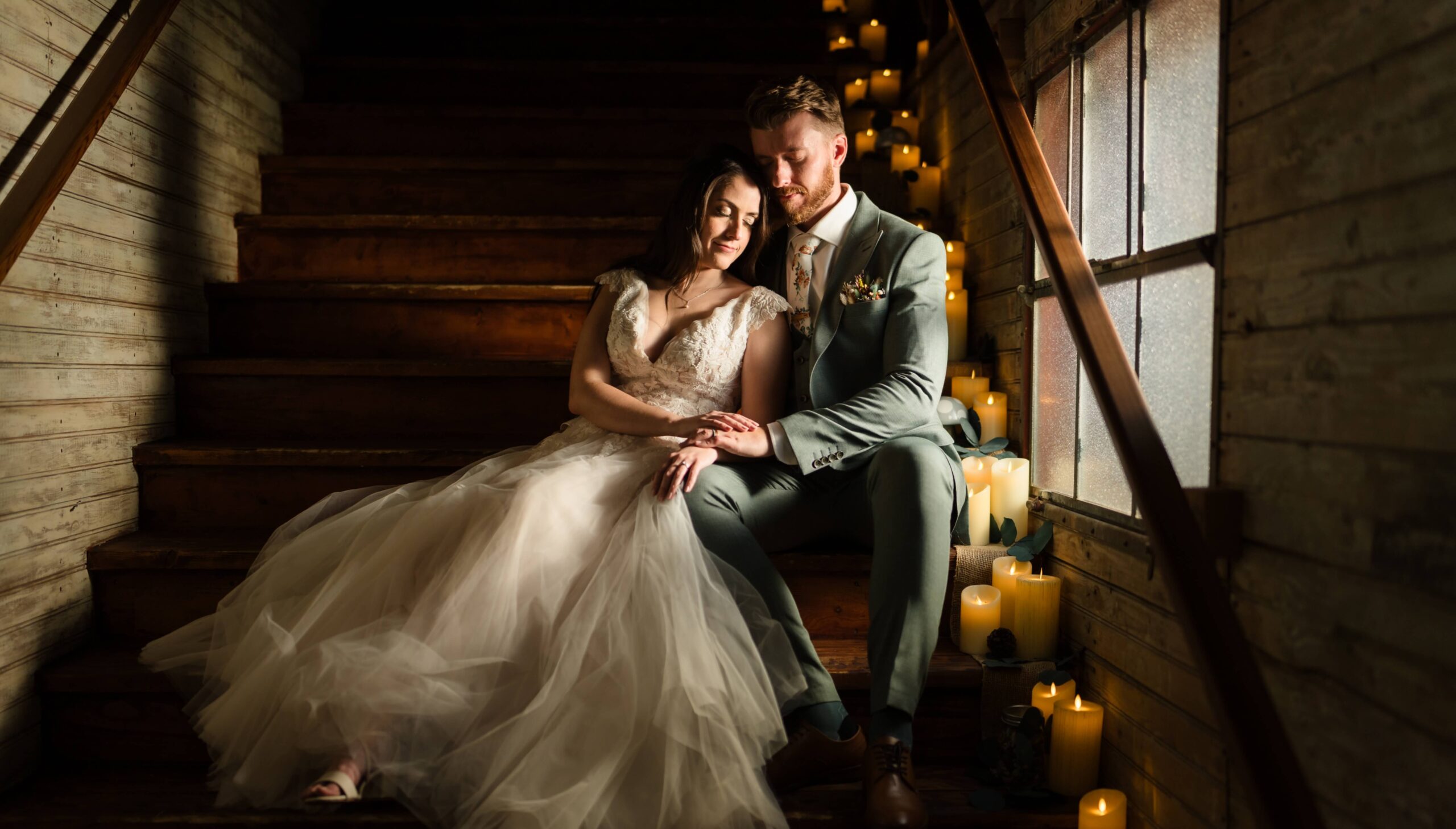A bride and groom sit together on wooden stairs, surrounded by lit candles. The bride wears a white gown, and the groom is in a light suit. They appear calm and content.