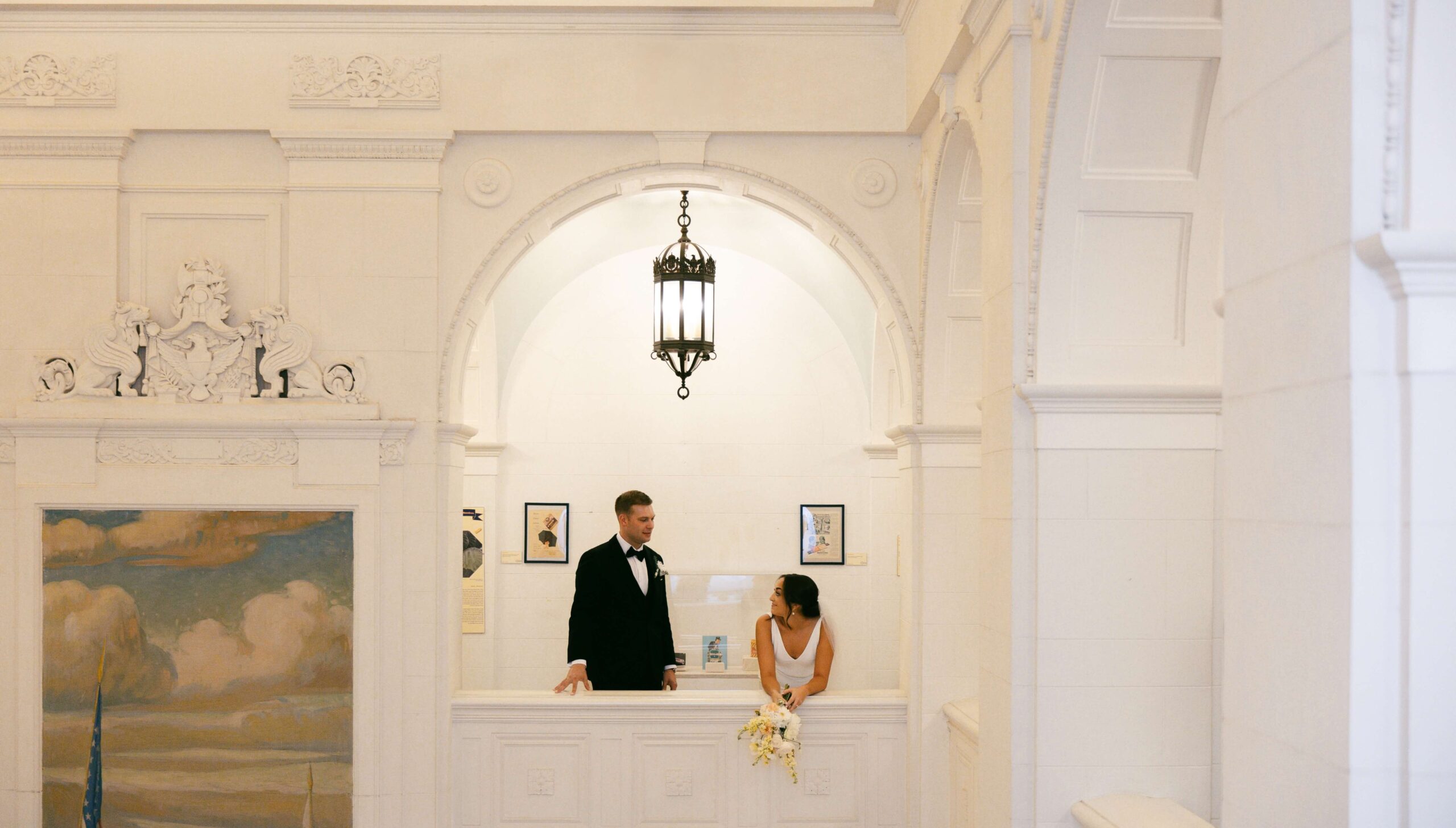 A bride and groom stand in an ornate white hallway, decorated with a chandelier and framed art, looking at each other. The bride holds a bouquet.