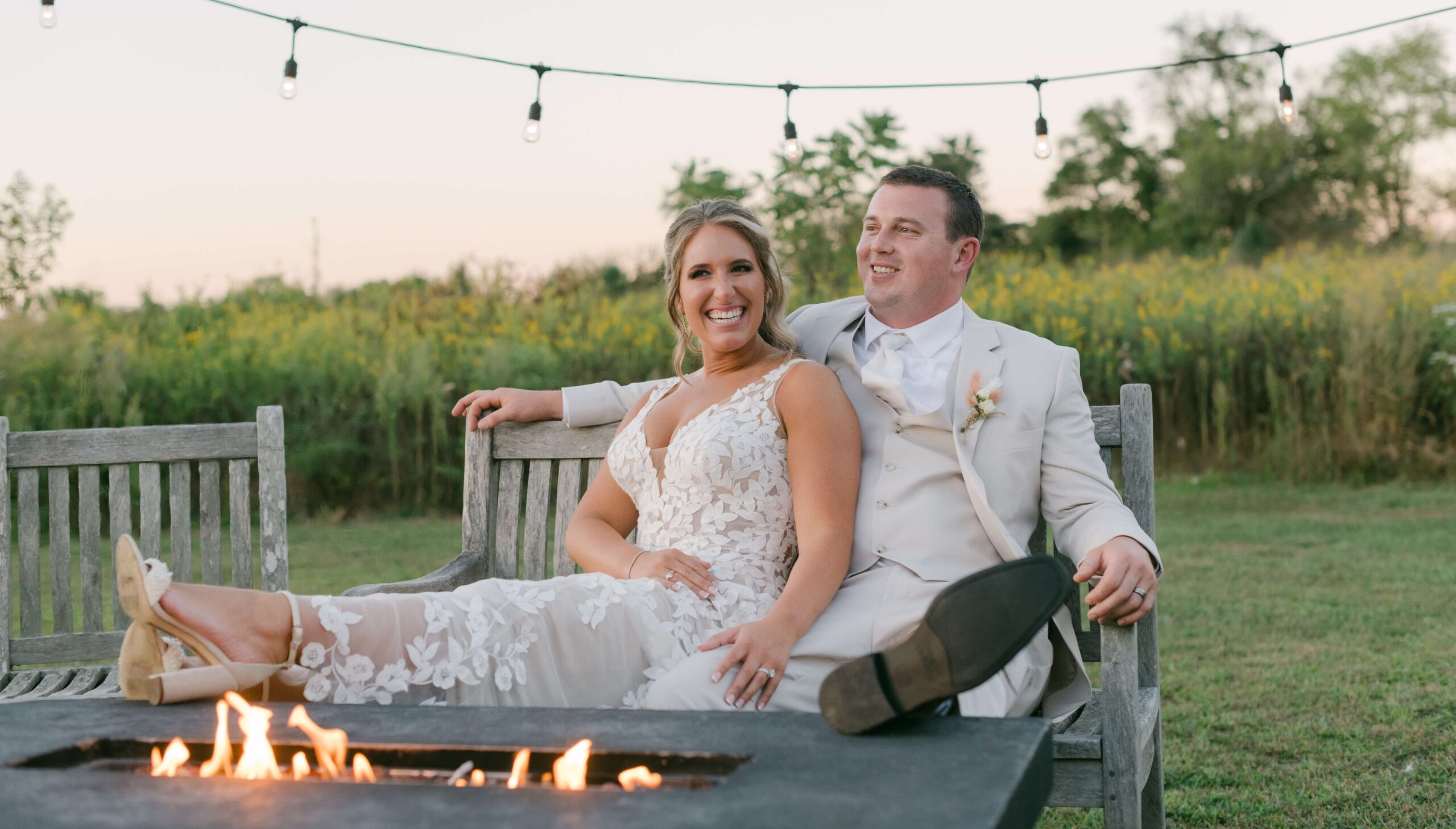 Bride and groom in wedding attire sit smiling on an outdoor wooden bench near a fire pit, with string lights and greenery in the background.