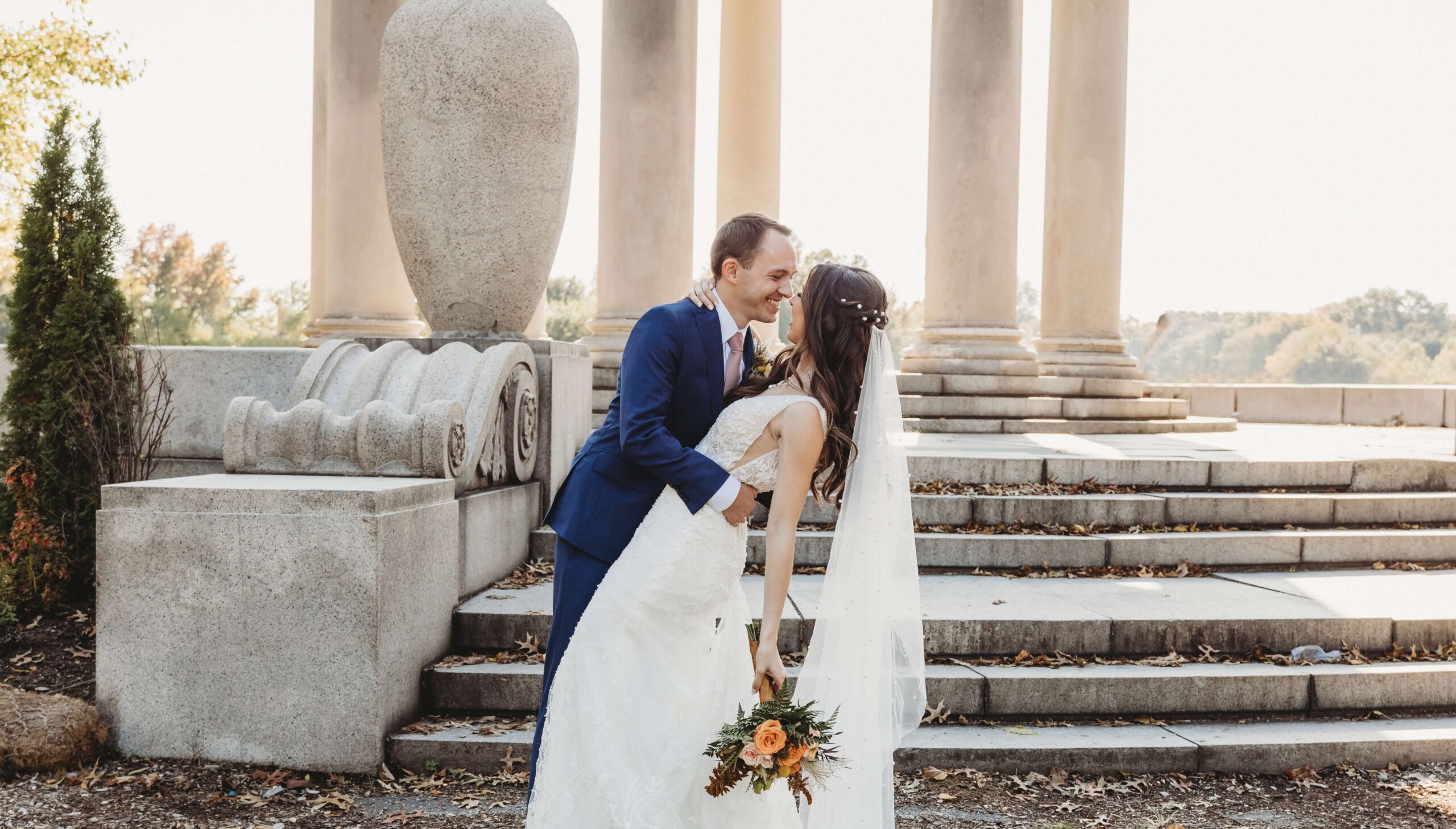 A groom in a blue suit kisses a bride in a white gown holding a bouquet near stone columns and steps.