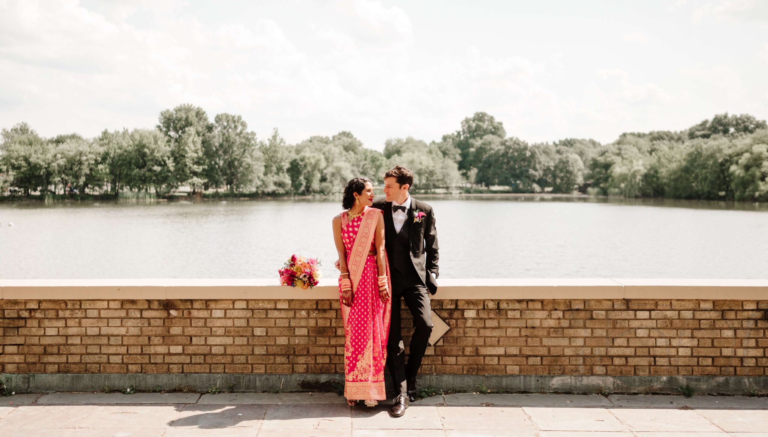 A couple stands by a brick wall overlooking a lake. The woman is in a red sari and the man in a black suit. A bouquet rests on the wall beside them. Trees are visible in the background.
